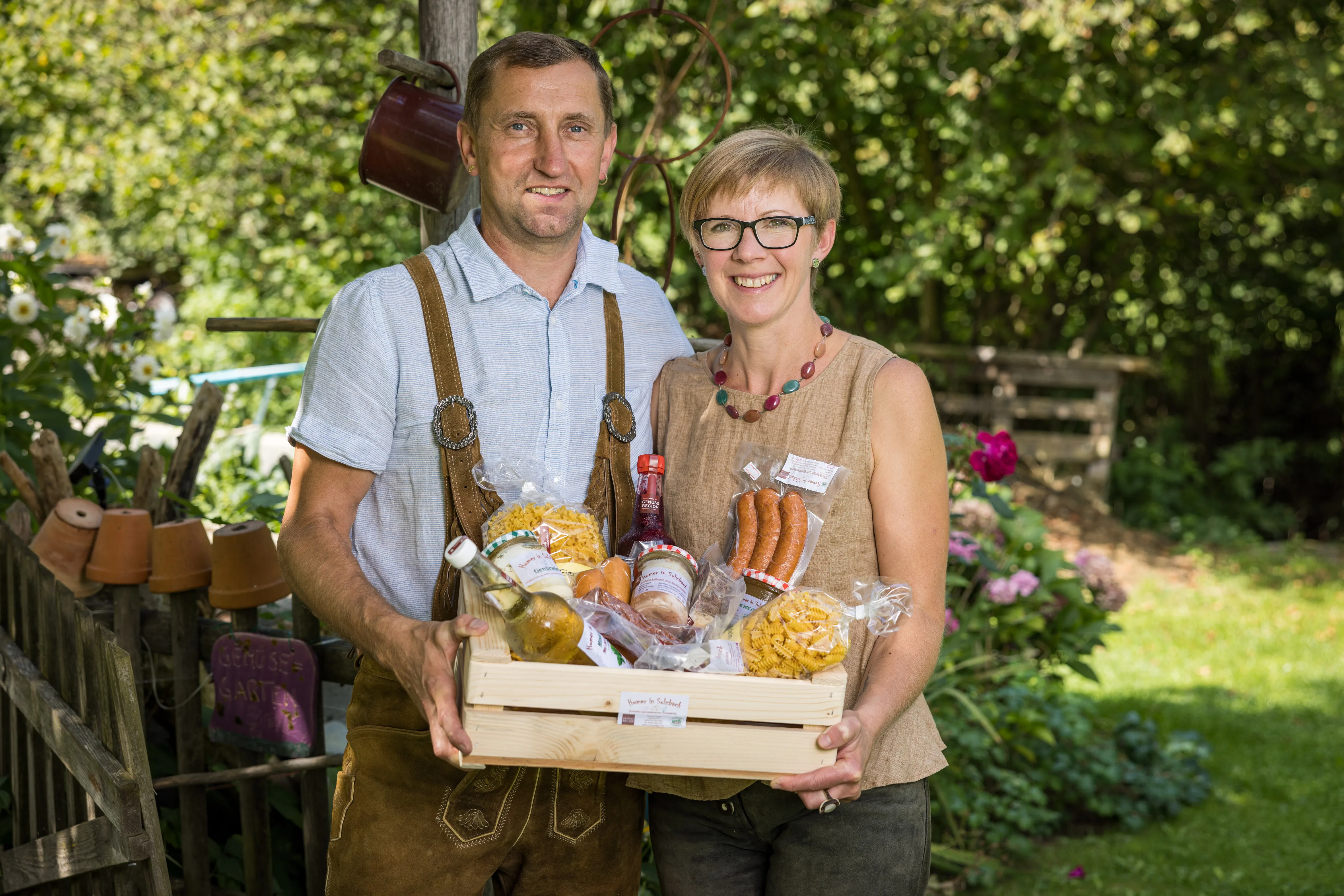 Ein Man und eine Frau mit Brille halten eine Kiste mit Produkten wie Nudeln, Würste, .. in den Händen. Dahinter grüne Wiese und Bäume und Sträucher