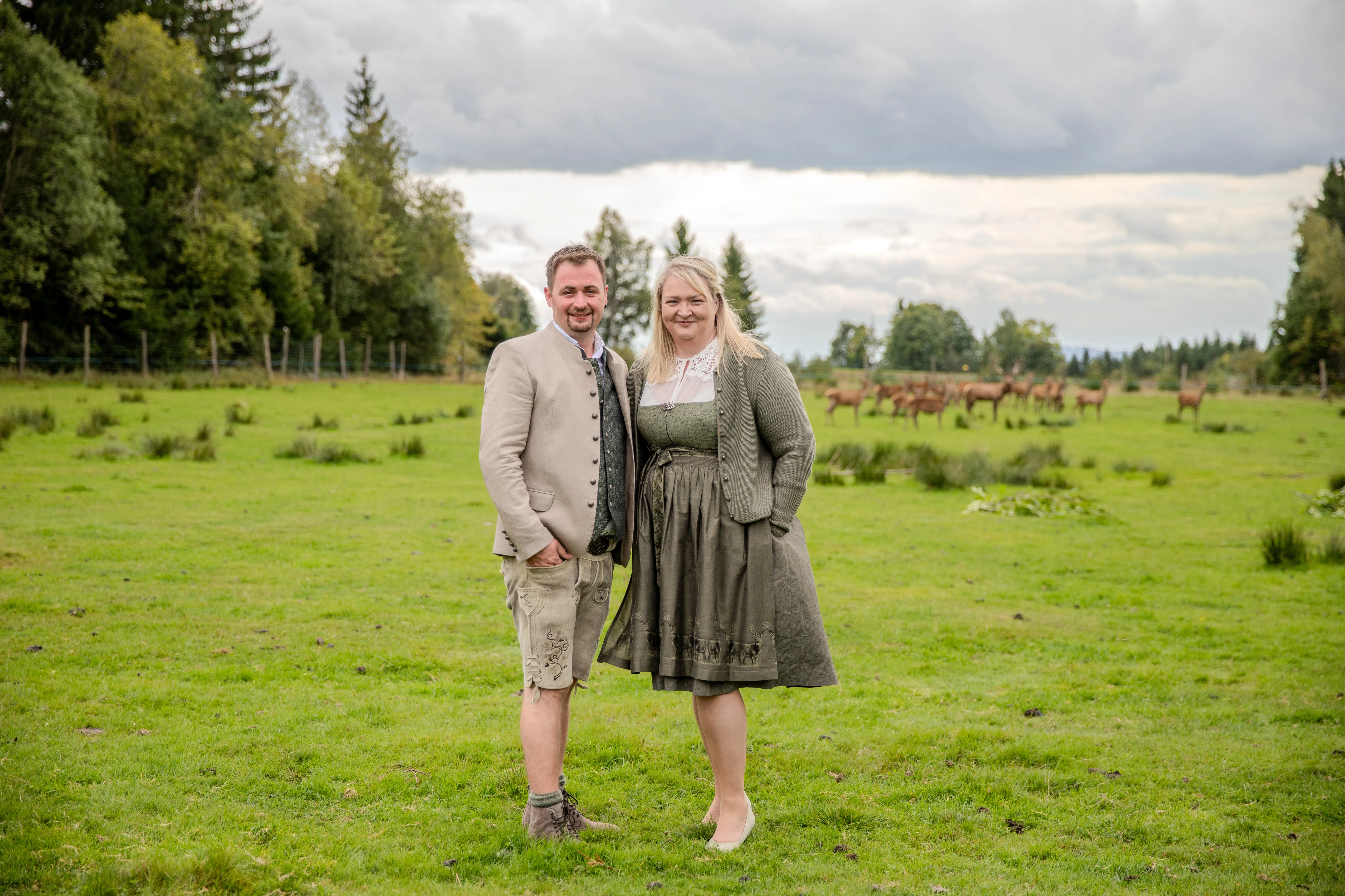 Eine Frau im Dirndl und ein Mann im Lederhose mit Trachtenjanker stehen auf einer Wiese; im Hintergrund ist Rotwild zu sehen und seitlich ein Wald bzw. einzelne Bäume, dahinter Himmel mit dunklen Wolken