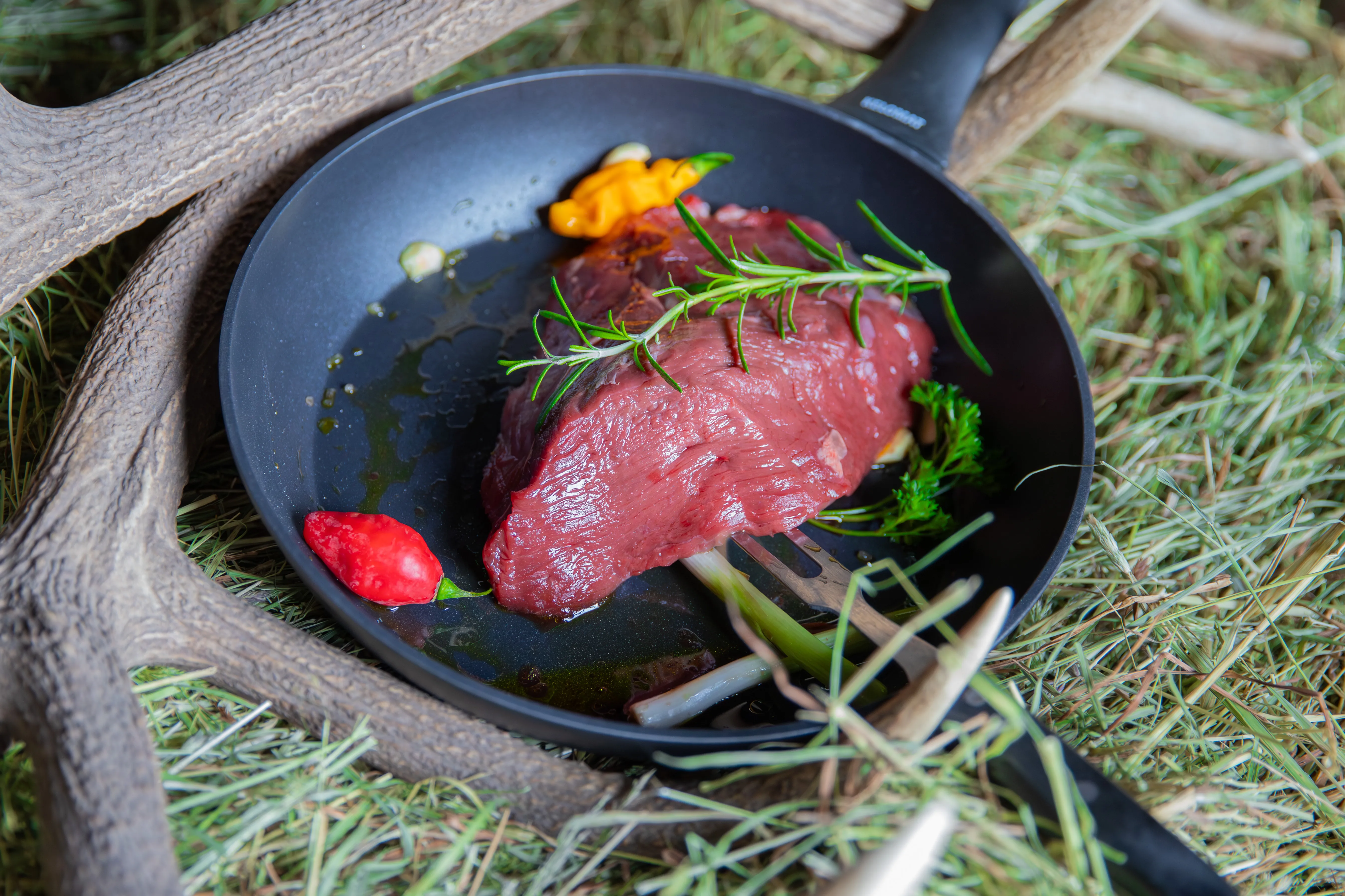 Eine schwarze Pfanne mit Frischfleisch und Kräutern steht auf Heu, daneben ein Hirschgeweih