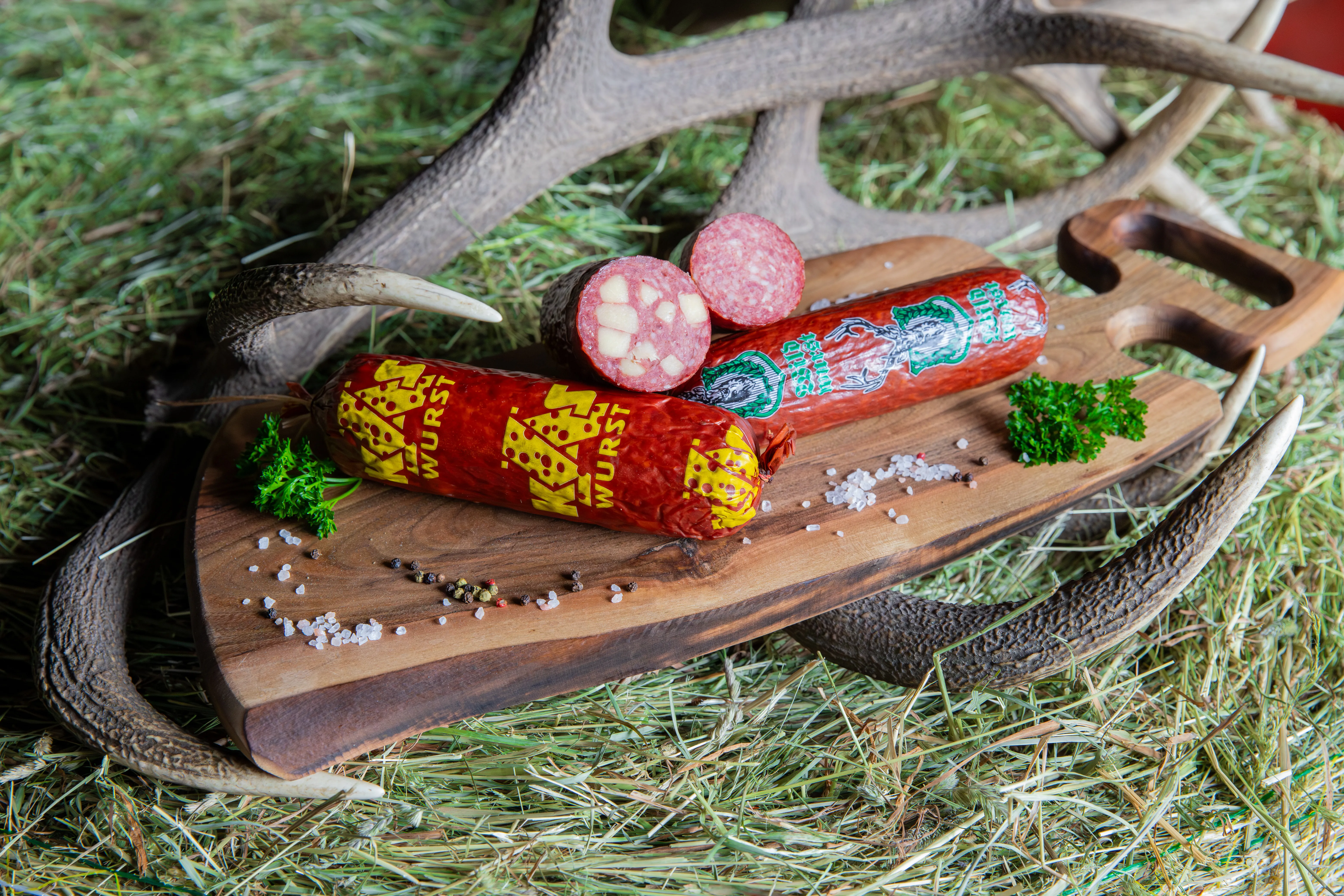 Ein Holzbrett mit Wurst im Ganzen inkl. Kräuter und Salz- bzw. Pfefferkörner liegt in einem Hirschgeweih im Heu.