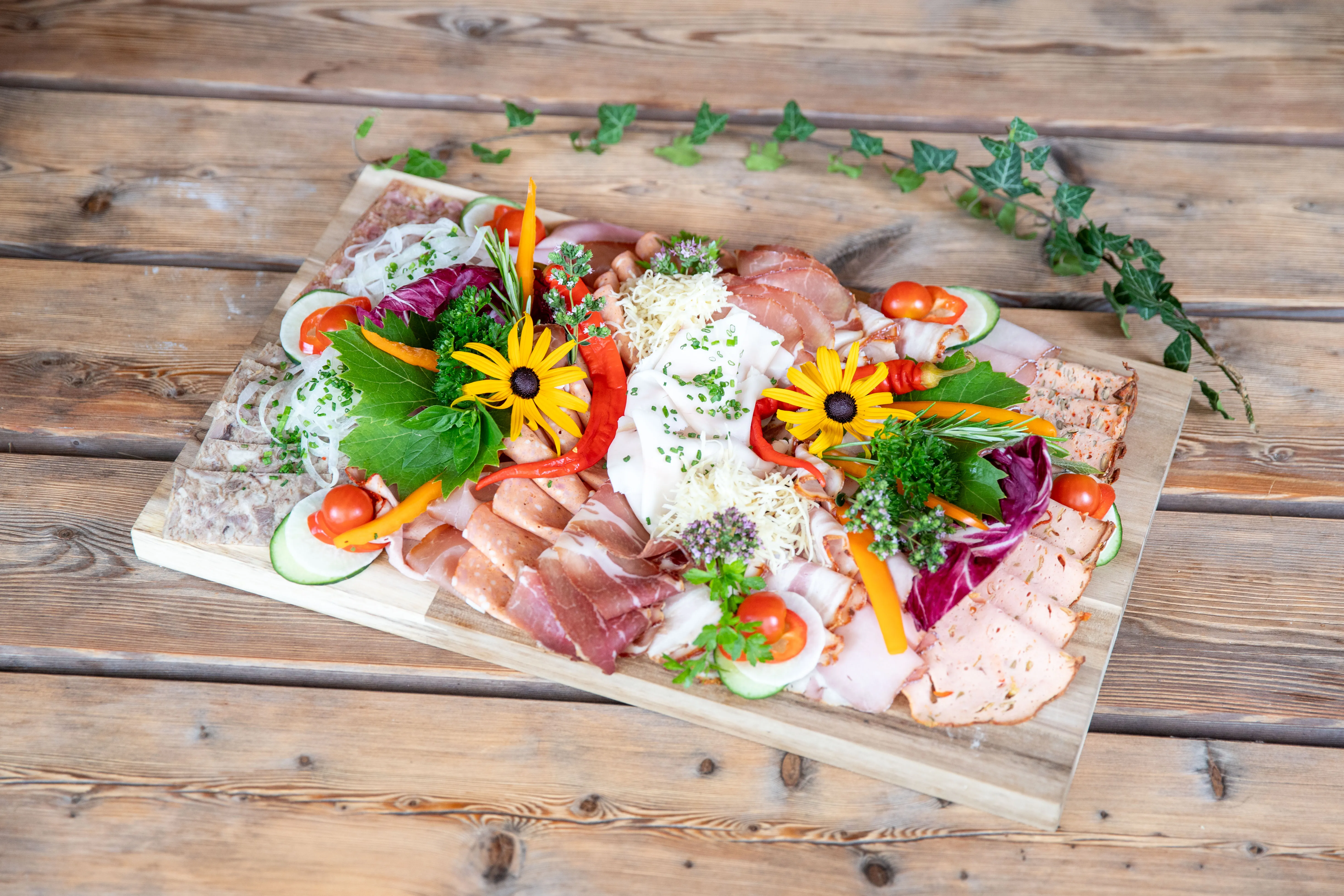 Auf einem Holztisch steht ein Holzbrett mit verschiedenen Fleisch- und Käseprodukten, garniert mit Kräuter, Eier und Blüten.