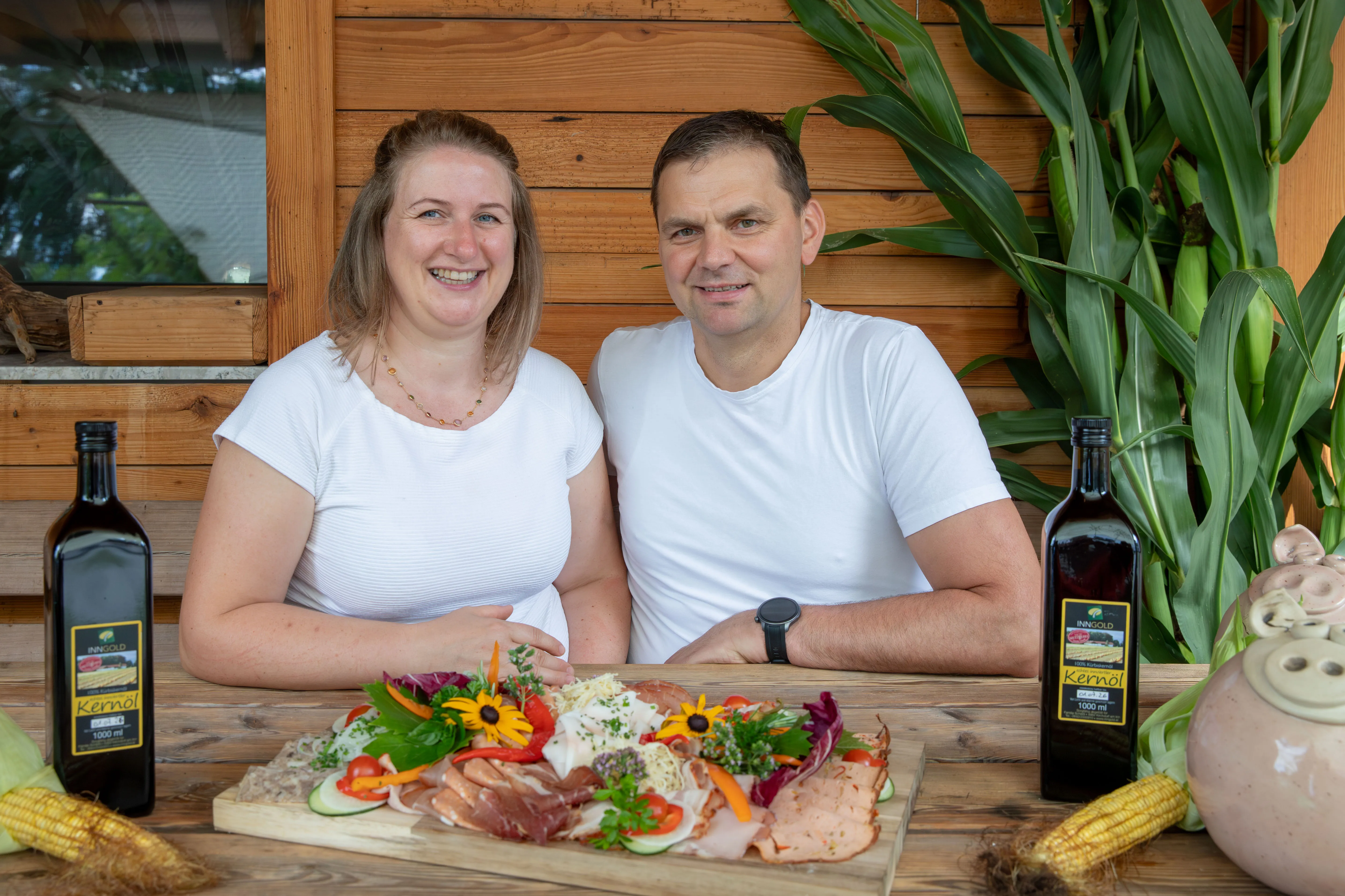 Ein Man und eine Frau im weißen T-Shirt sitzen an einem Tisch; davor ein großes Holzbrett mit vielen verschiedenen Fleischprodukten; daneben zwei Flaschen mit Kernöl und Maiskolben. Dahinter eine Holzwand und eine Grünpflanze