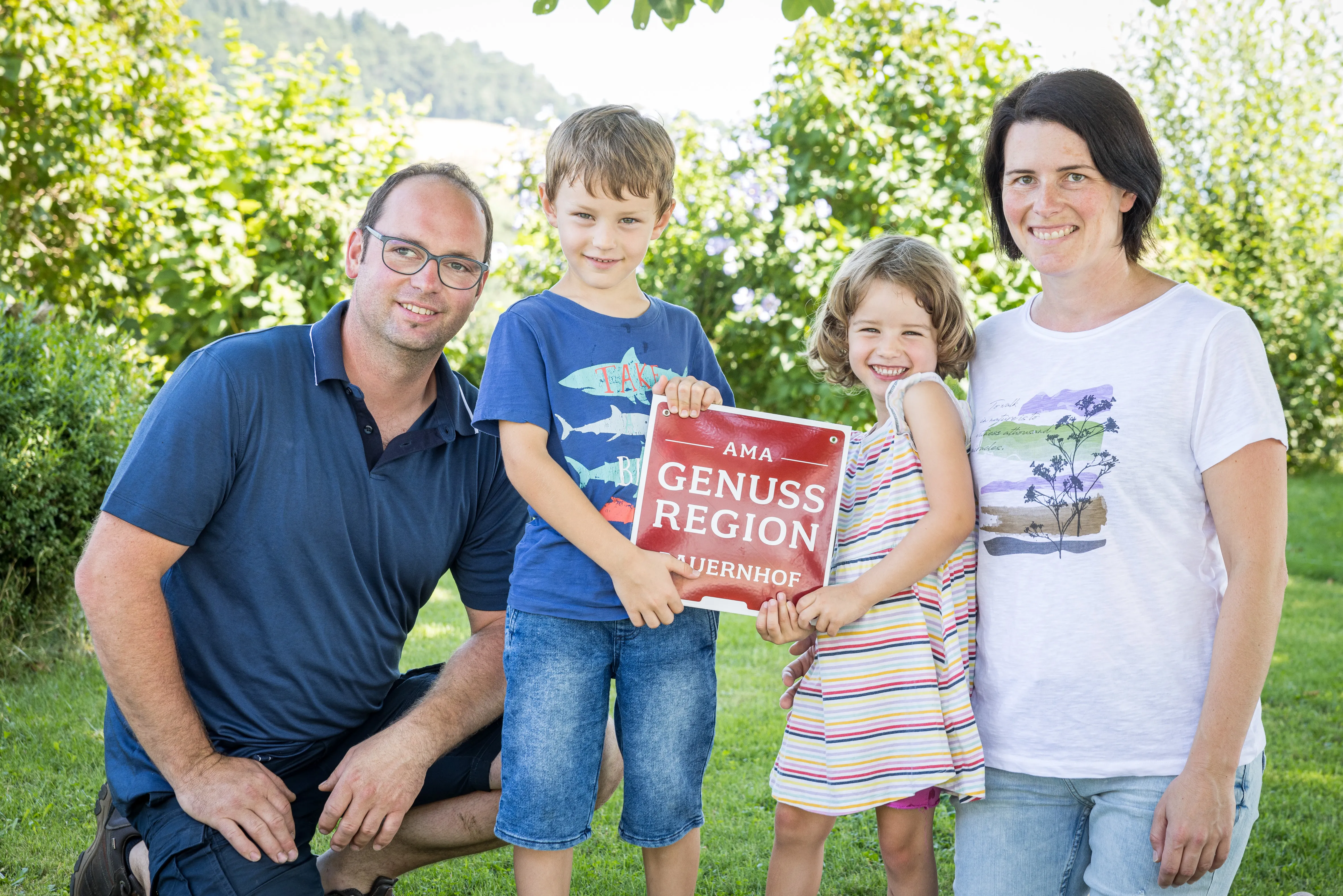 Herr und Frau Kagerer knieen mit ihren zwei Kindern auf einer Wiese. Dahinter grüne Sträucher