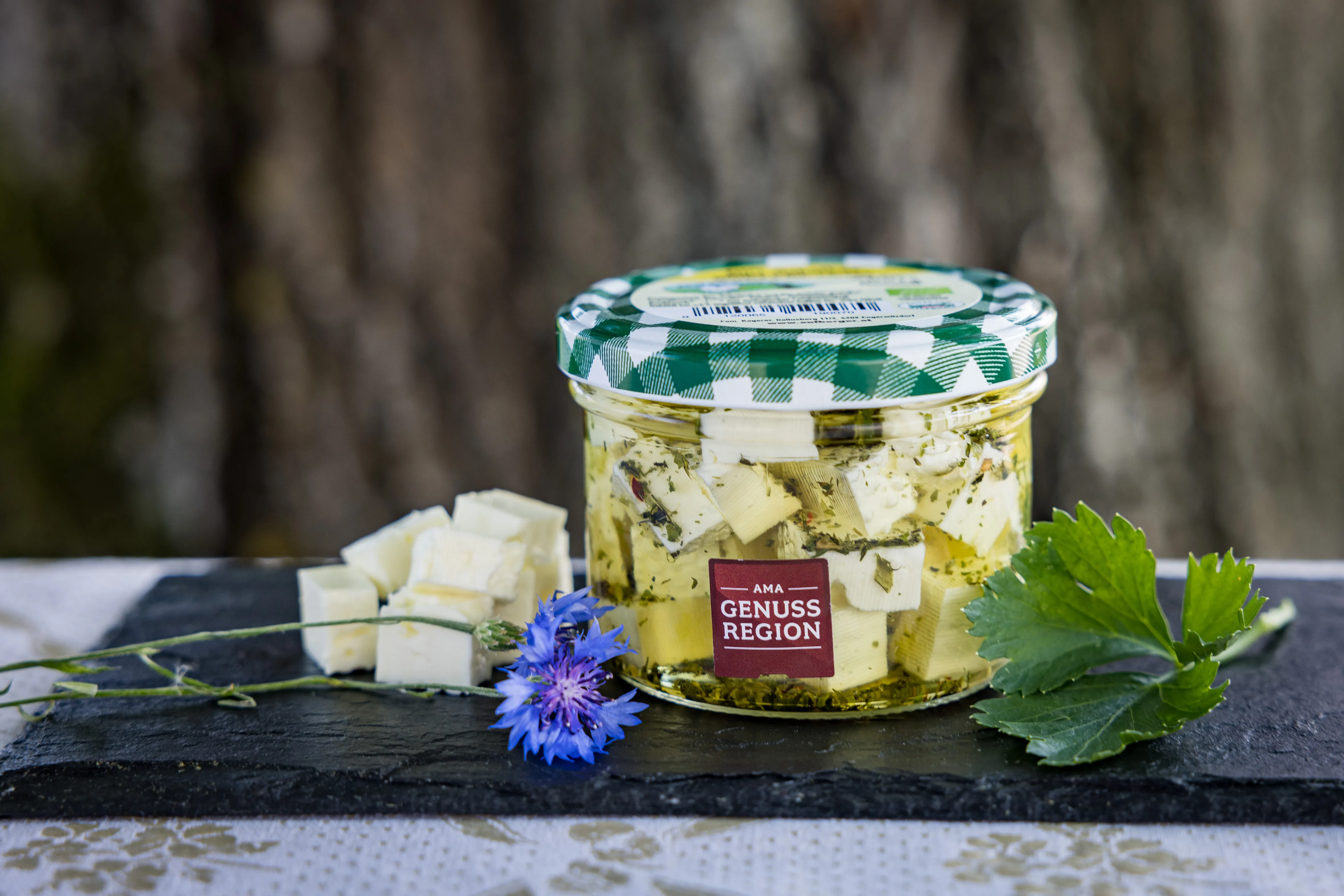 Auf einer schwarzen Schieferplatte steht ein Glas mit eingelegtem Käse, daneben einige Käsestückchen extra. Garniert mit einer blauen Kornblume und Blätter.