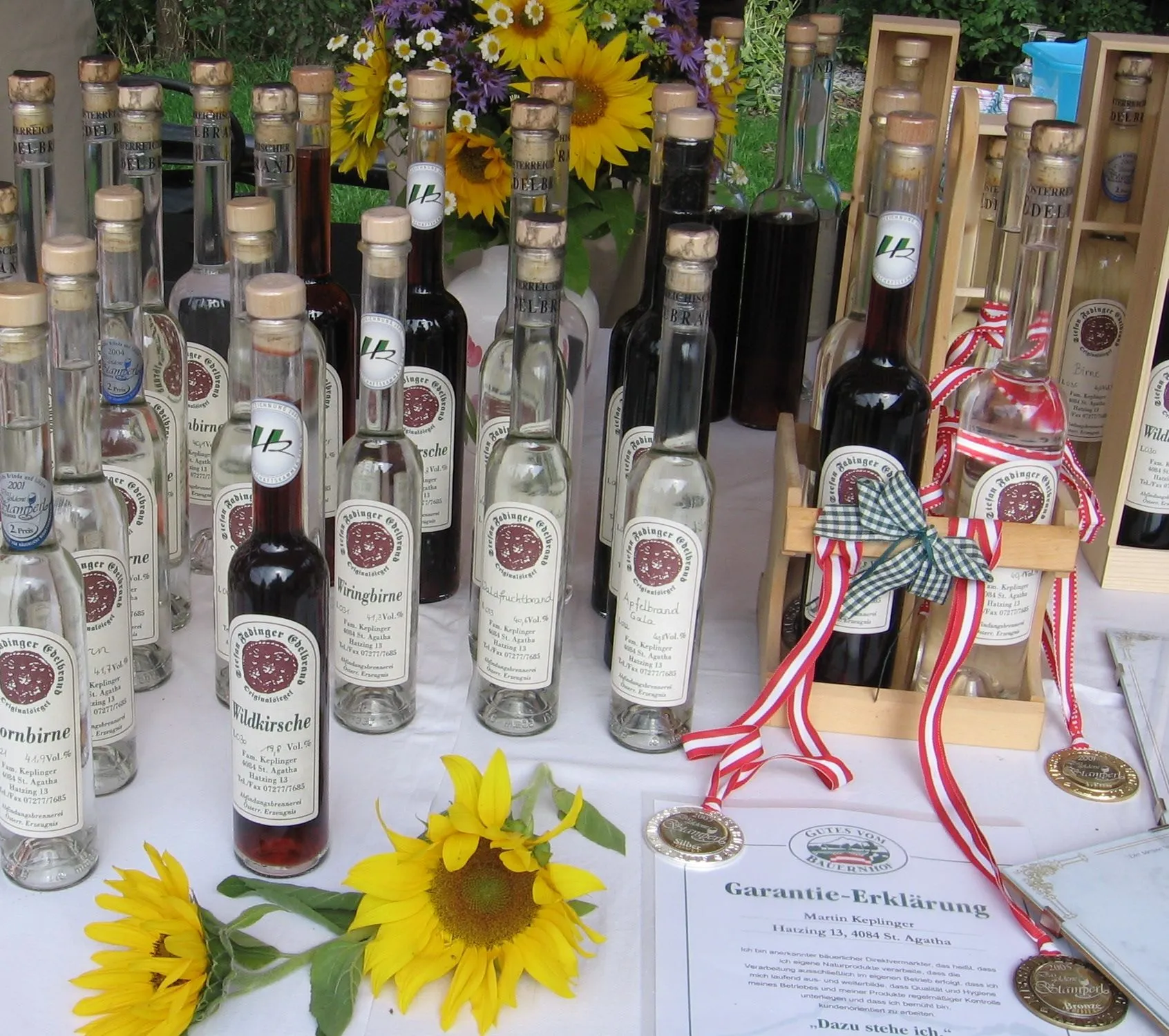 Auf einem Tisch mit weißem Tuch stehen viele Flaschen verschiedener Brände und Liköre, daneben Medaillen und Urkunde, dazwischen Sonnenblumen