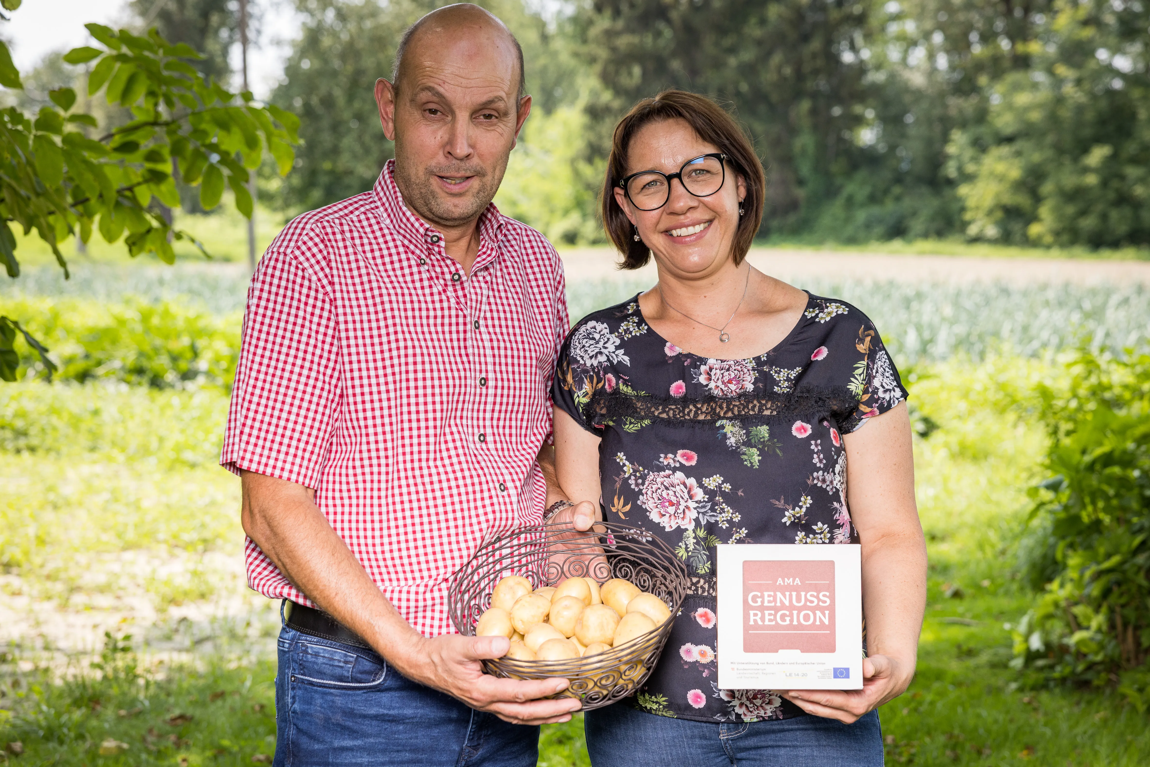 Herr und Frau Kirchmayr stehen auf einer Wiese und halten einen Korb mit Kartoffel und eine kleine Tafel mit der Aufschrift AMA GENUSS REGION in den Händen. Dahinter grüne Wiesen, Felder und Wald.