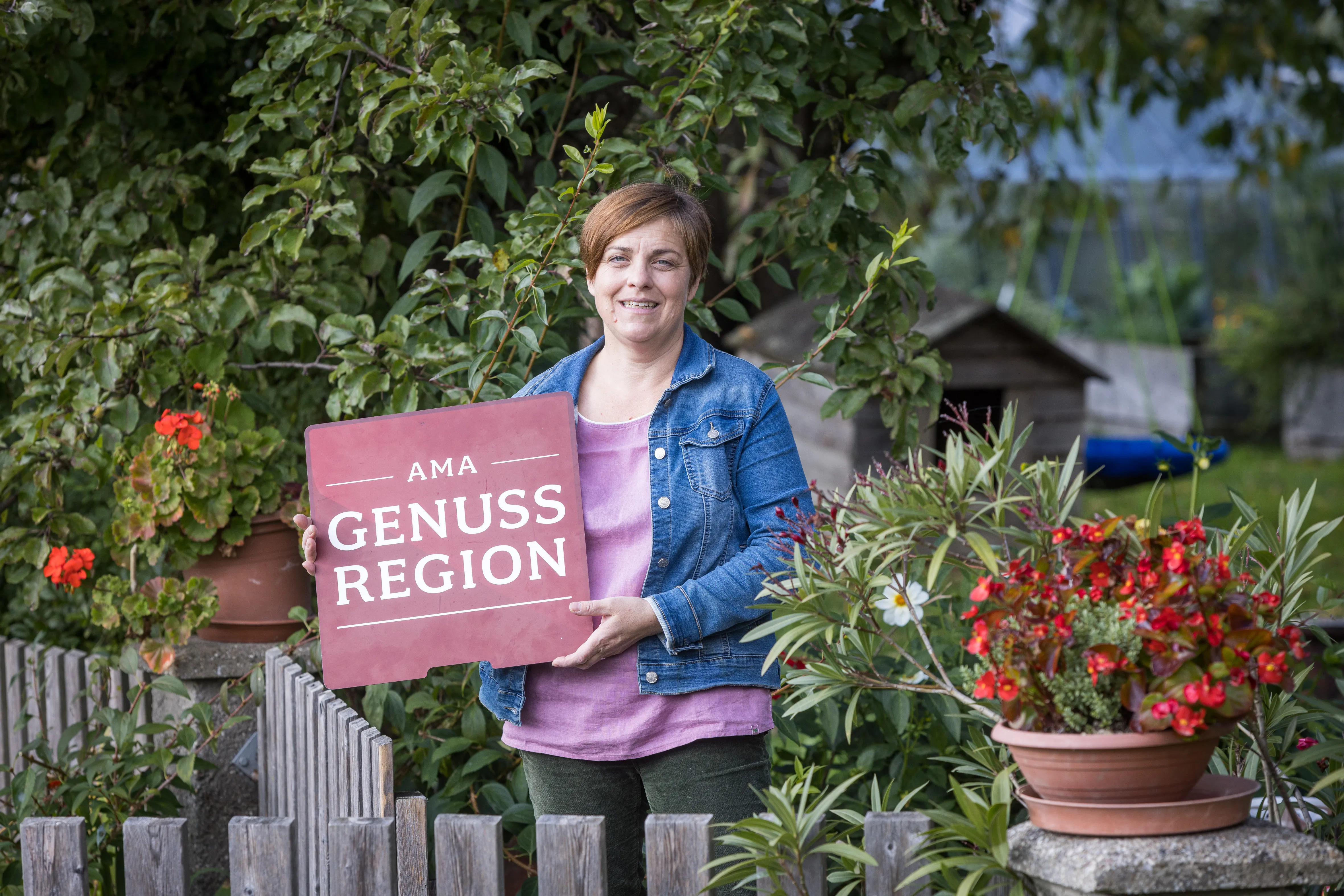 Eine Frau in Jeansjacke hält eine Tafel mit der Aufschrift Ama Genuss Region in der Hand und steht in einem Garten; daneben ein Holzzaun und rote blühende Blumen, dahinter noch Bäume und Gebäude