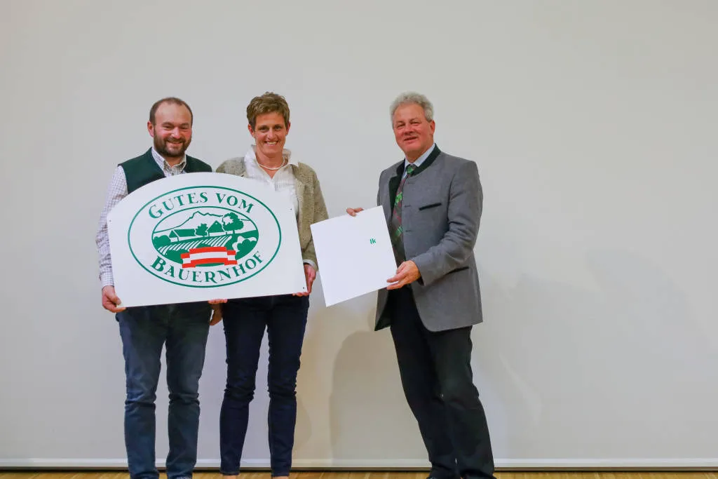 Herr und Frau Kreil stehen mit einem weiteren Mann vor einem weißen Hintergrund und halten eine Hoftafel und eine Urkunde in die Kamera
