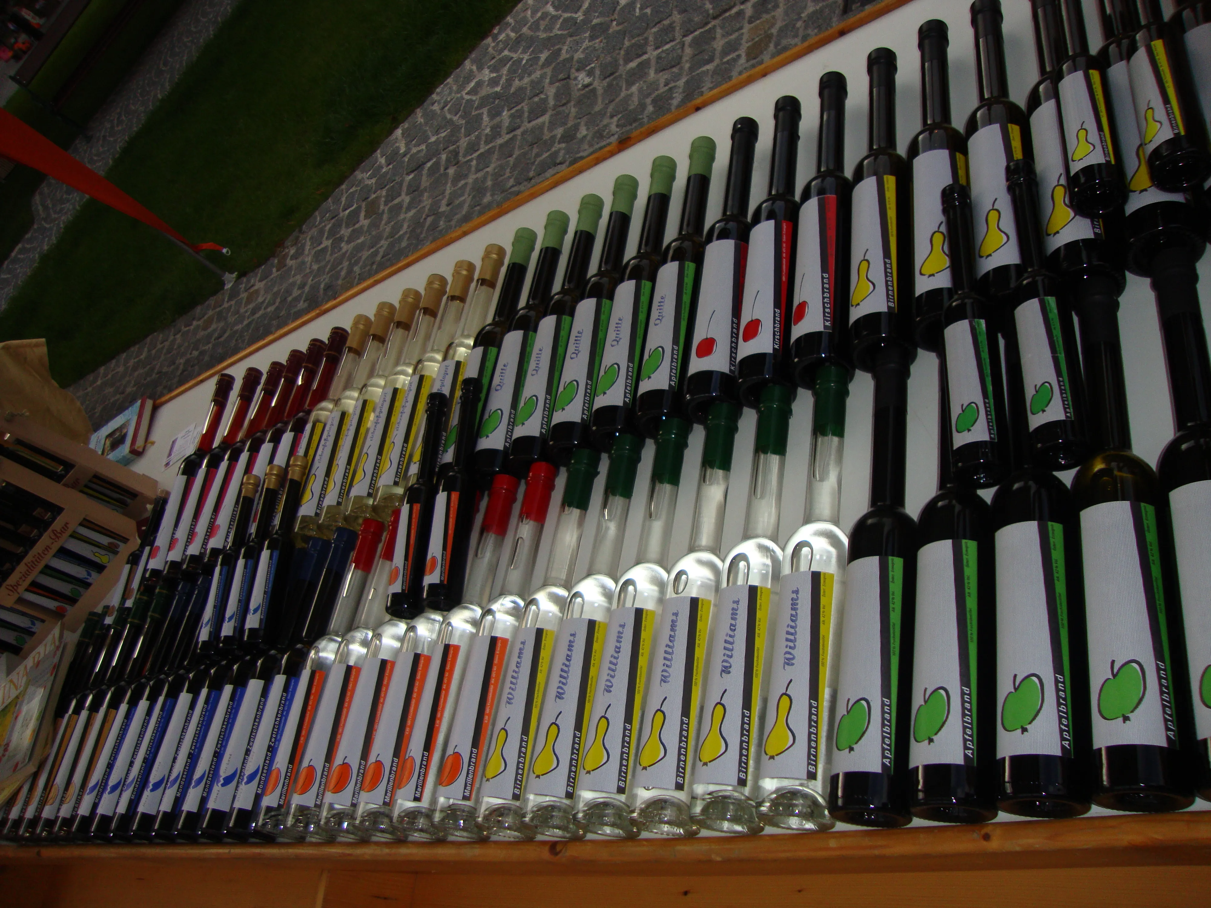 An einer Wand stehen mehrere Reihen Likör- bzw. Schnapsflaschen übereinander; verschieden färbige Etiketten, dahinter Steinmauer