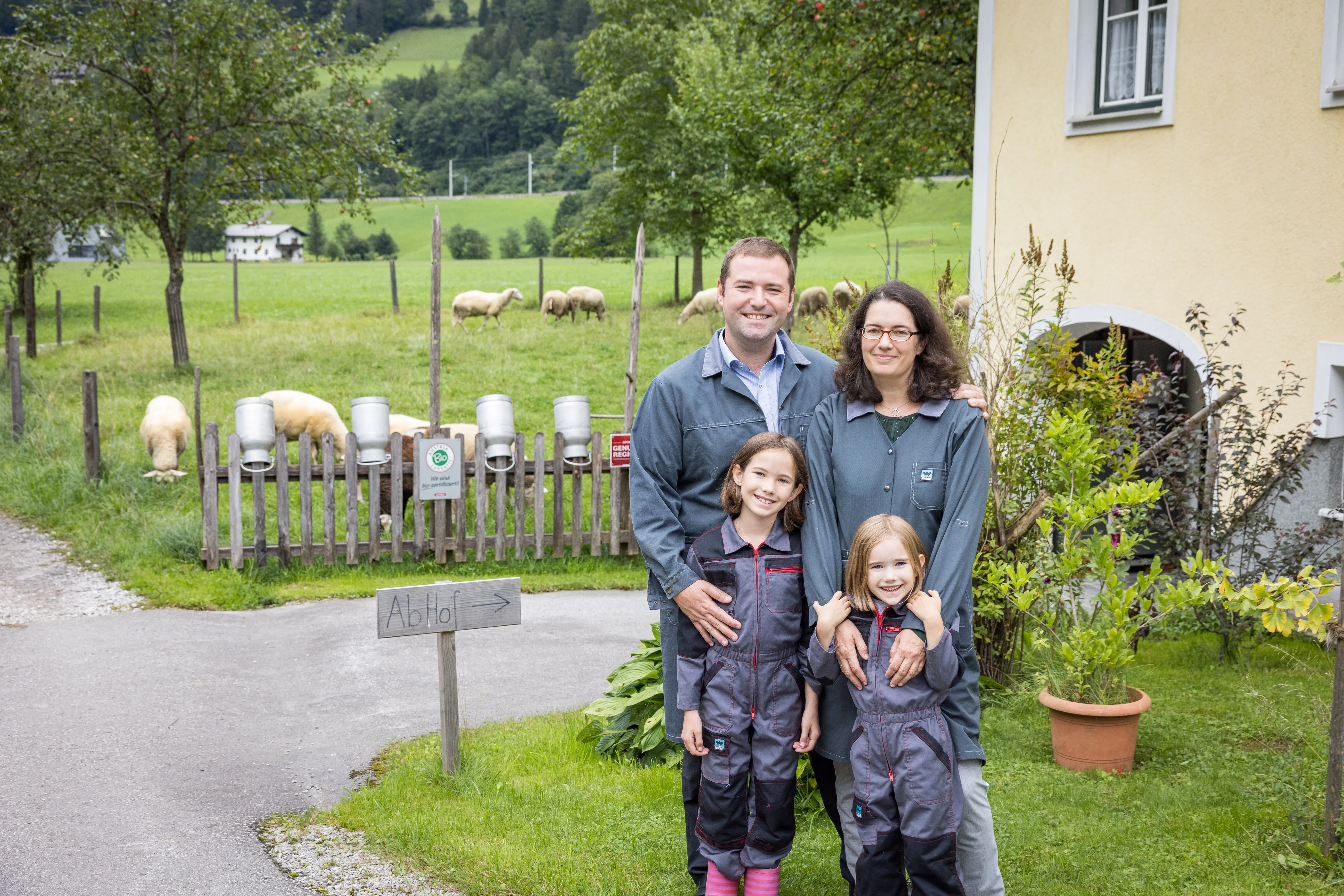 Ein Mann, eine Frau und zwei Mädchen stehen vor einem gelben Bauernhaus, daneben eine Koppel mit Schafen und viel Wiese und Bäume. Auf dem Zaun stecken mehrere große Milchkannen.