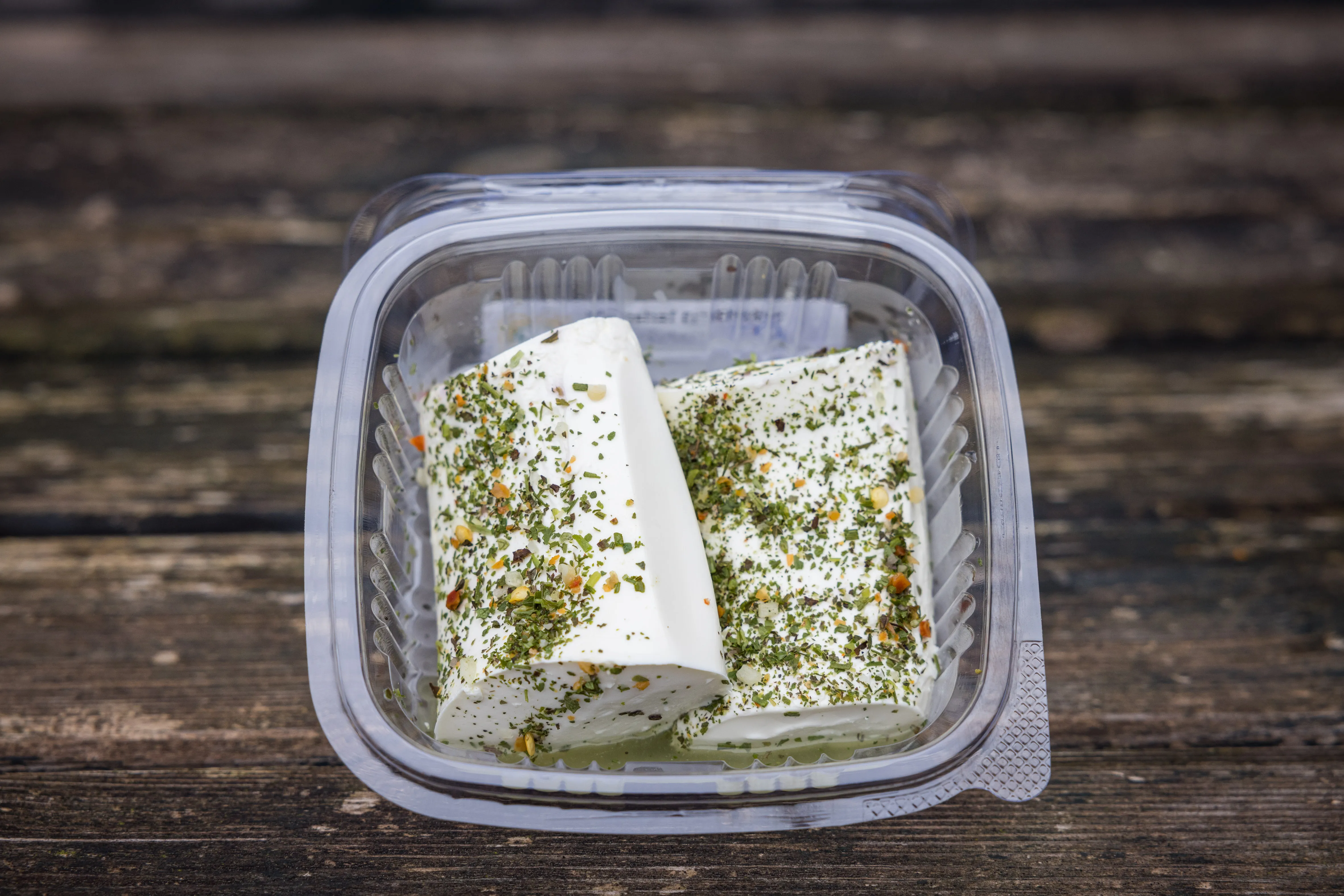 Auf einem dunklen Holzuntergrund liegt ein Plastikbehälter mit einem weißen Schafkäse, bestreut mit Kräutern.