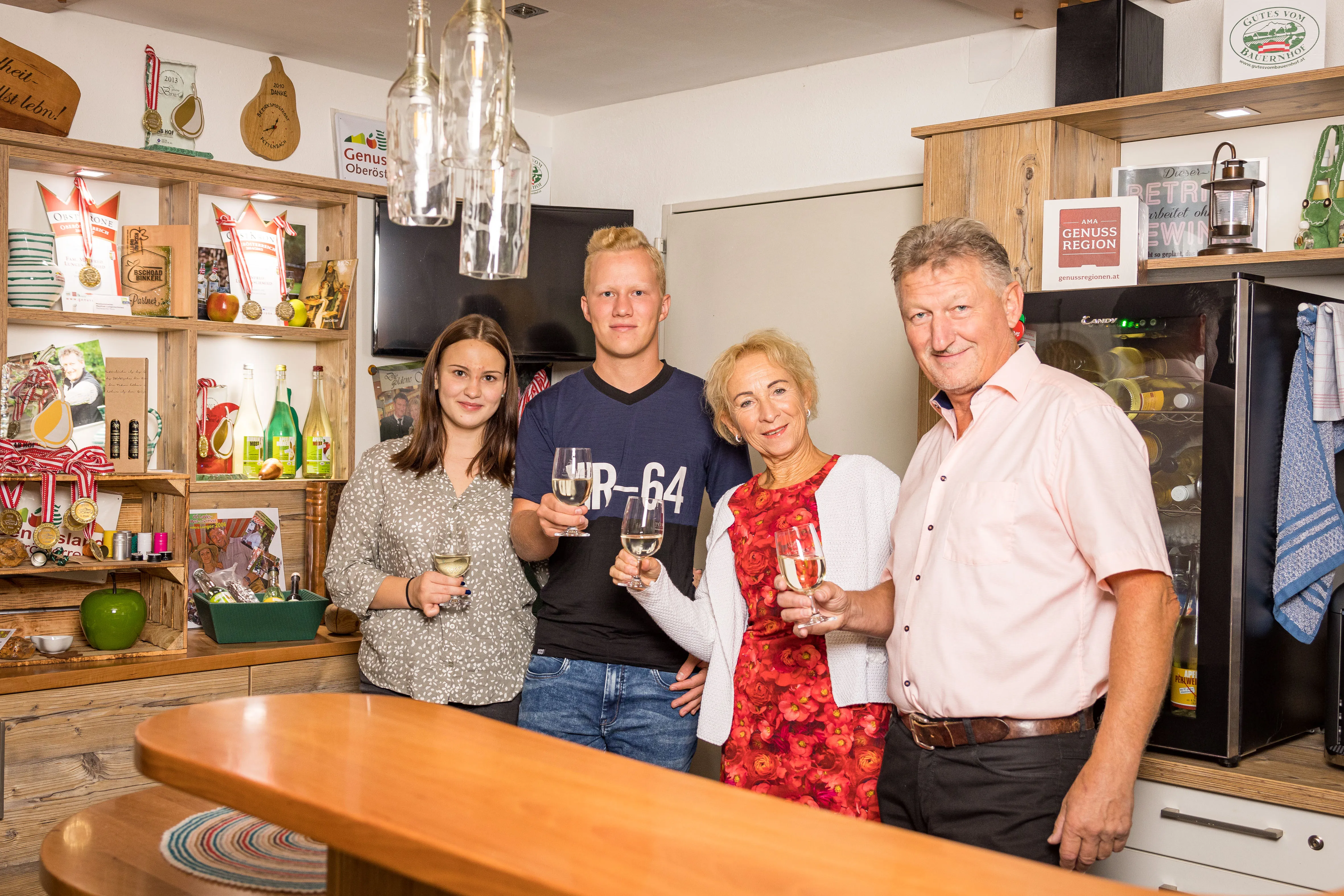Zwei Männer und zwei Frauen stehen mit einem Weinglas in einem Hofladen. Hinter ihnen Regale an der Wand mit verschiedenen Produkten