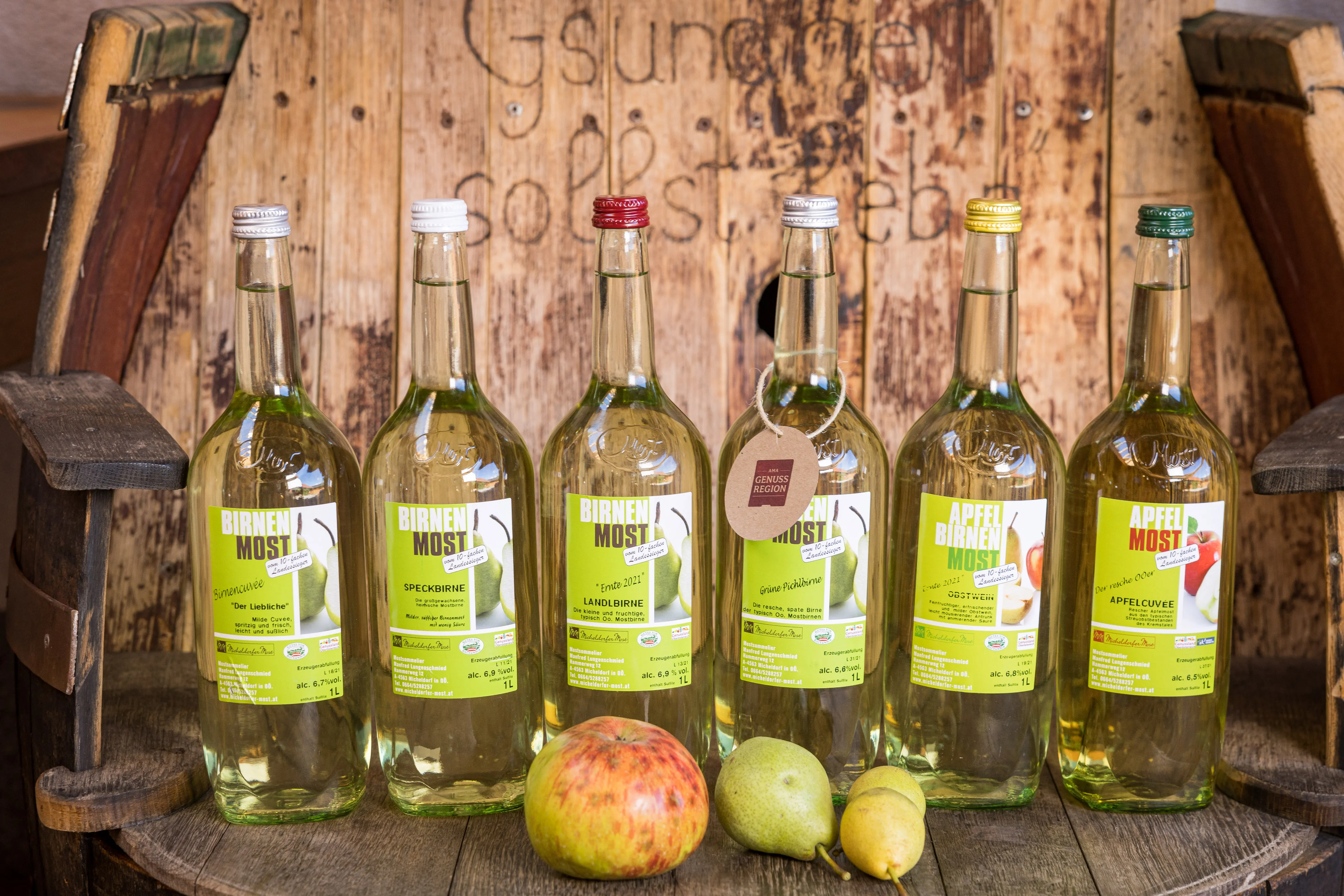 Auf einem Holzfass stehen mehrere Mostflaschen, davor liegen Äpfel und Birnen, dahinter eine Holzwand mit Spruch