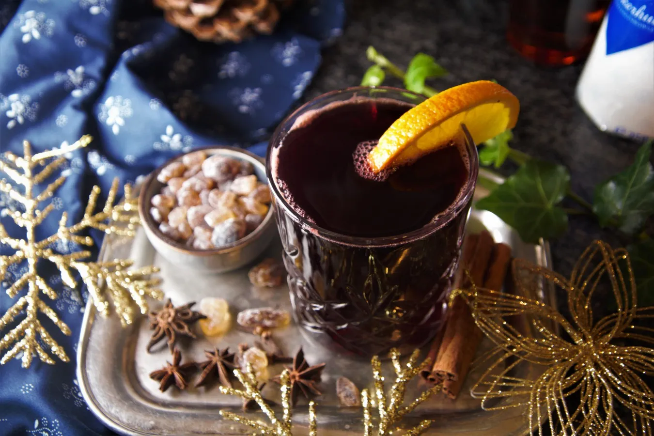 Glas mit Rotwein-Feuerzangenbowle, garniert mit Orangenscheibe, auf Tablett mit Zimtstangen, Sternanis, kandiertem Ingwer und goldfarbenen Deko-Schneeflocken.