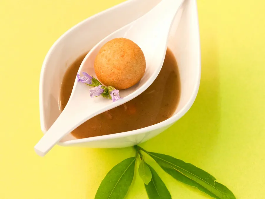 Weiße Schale mit brauner Suppe, darauf ein weißer Löffel mit einem runden Gebäck und kleinen lila Blüten, grüne Blätter liegen darunter.