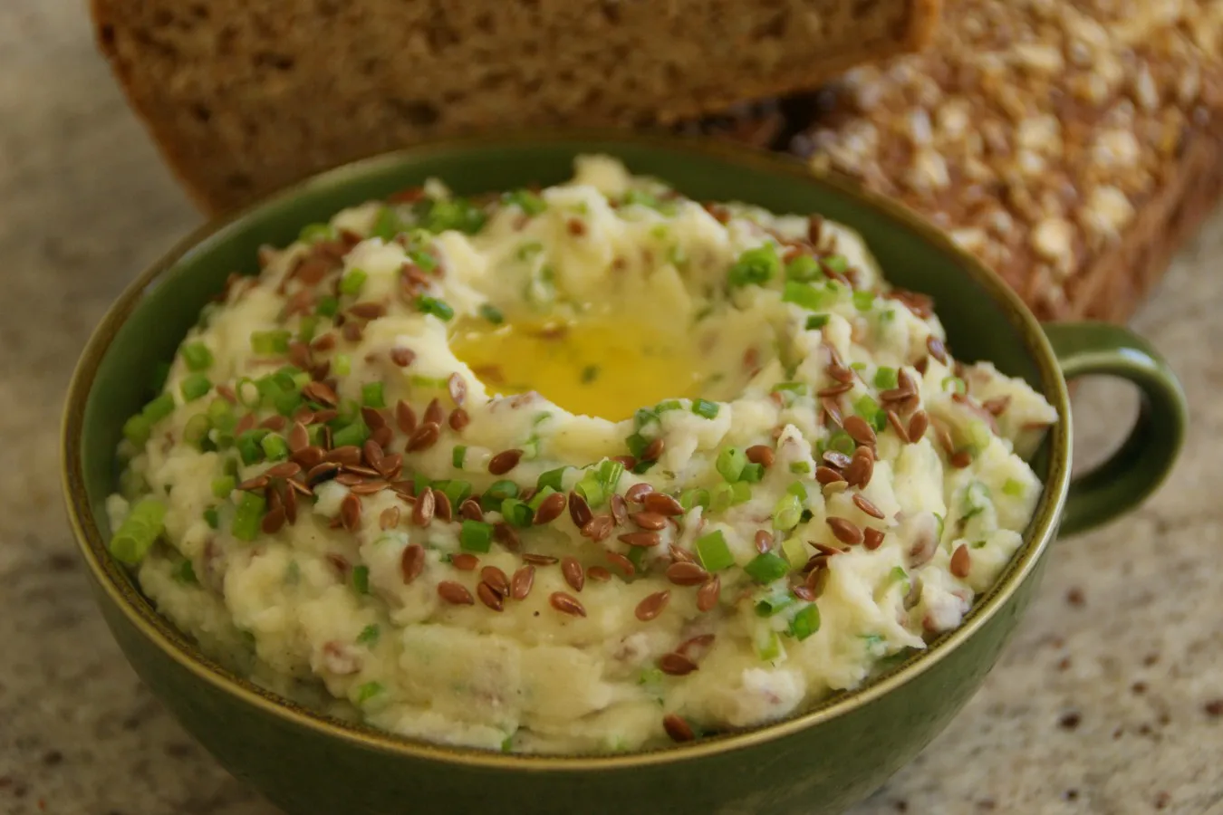 Grüne Schale mit cremigem Erdäpfel-Leinölaufstrich, bestreut mit Leinsamen und Schnittlauch, im Hintergrund zwei Scheiben Vollkornbrot.