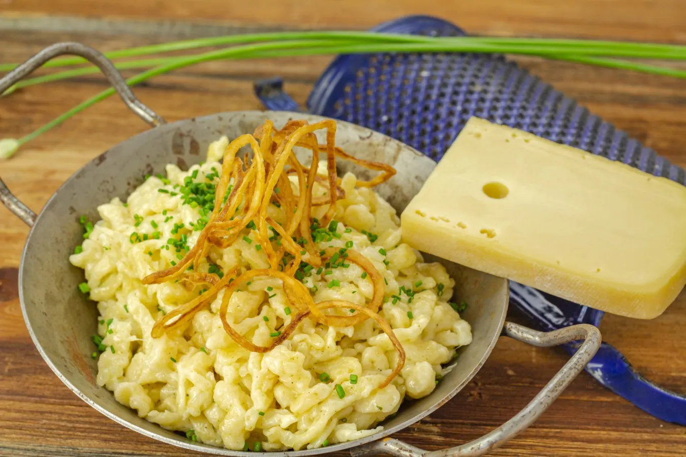 In einer Eisenpfanne befinden sich hellgelbe Spätzle, garniert mit Käse und, goldbraun gebratenen Zwiebelringen und Kräutern.