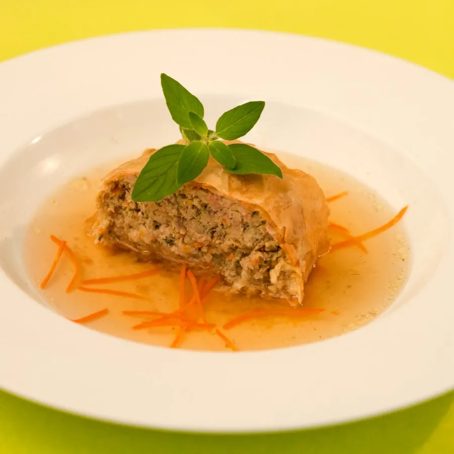 Gefüllter Fleischstrudel in klarer Brühe mit Karottenstreifen, garniert mit frischen Basilikumblättern auf weißem Teller.