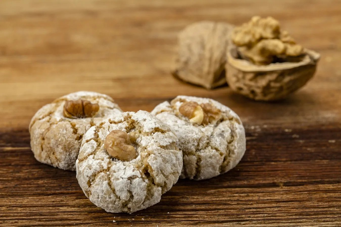 Drei runde, mit Puderzucker bestäubte Nusserlbusserl mit Walnussstücken darauf, im Hintergrund eine geöffnete und eine geschlossene Walnussschale auf Holzuntergrund.