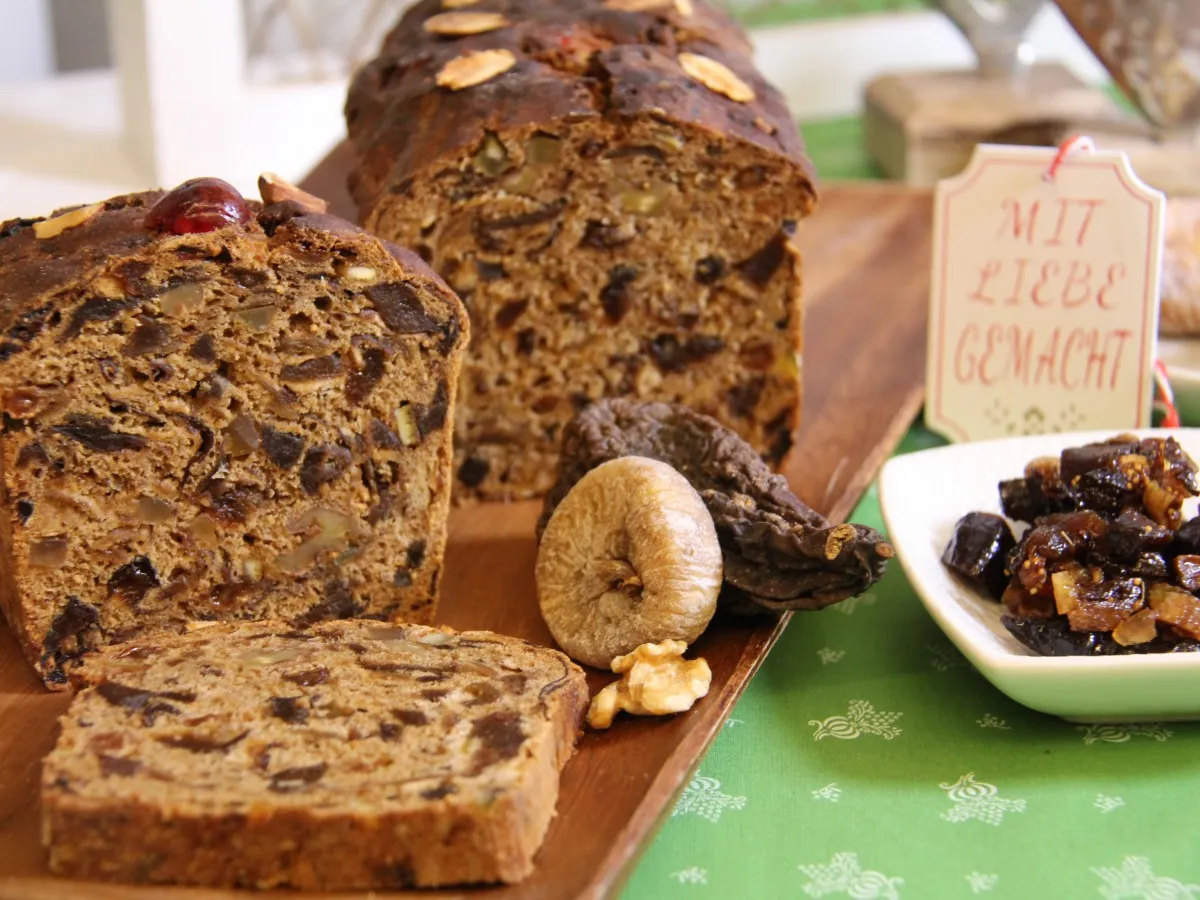 Braunes Früchtebrot mit Nüssen und Trockenfrüchten, angeschnitten auf Holzbrett, daneben getrocknete Feigen und Walnüsse, Schild mit roter Schrift 'MIT LIEBE GEMACHT'.