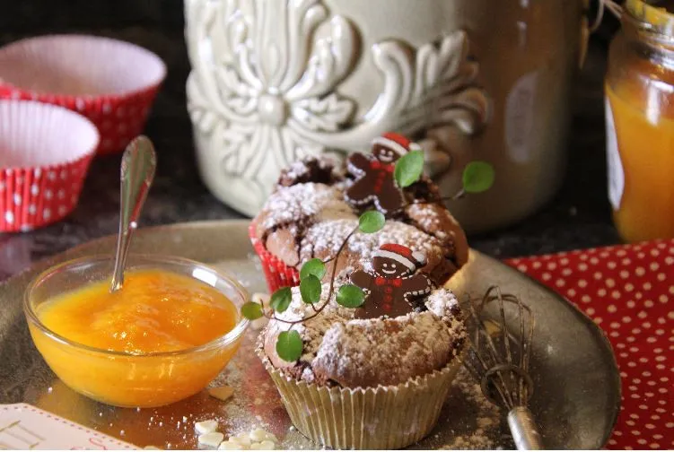 Zwei Muffins mit Puderzucker, verziert mit kleinen Lebkuchenmännchen und grünen Blättern, daneben eine Glasschale mit oranger Marmelade und ein kleiner Schneebesen.