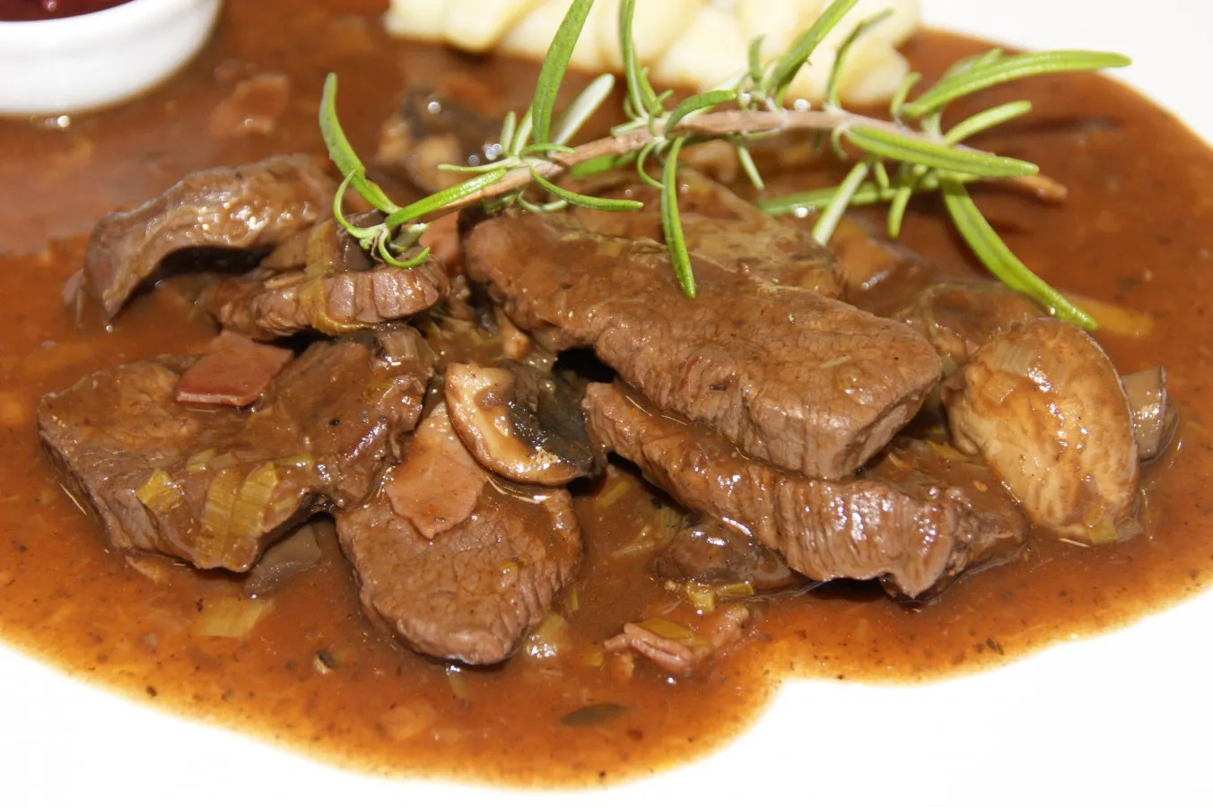Mehrere Scheiben Rindfleisch in brauner Sauce mit Pilzen und Rosmarinzweig als Garnitur.