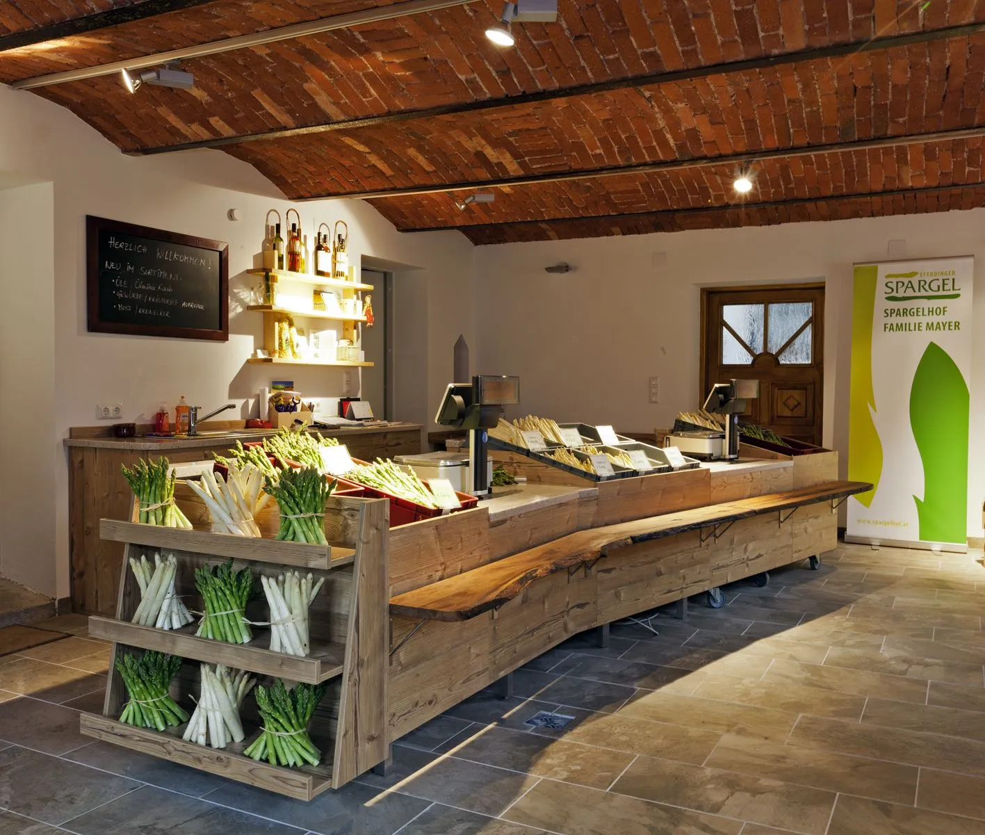 In einem Raum mit Holzdecke steht ein Holztresen mit frischem Spargel, dahinter eine Spüle und ein Regal an der Wand.