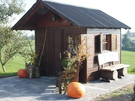 Auf einem gepflastertem Platz steht eine kleine Holzhütte mit einer Bank davor. zwei Kürbisse zur Zierde neben der Hütte, rundherum Wiesen und Bäume