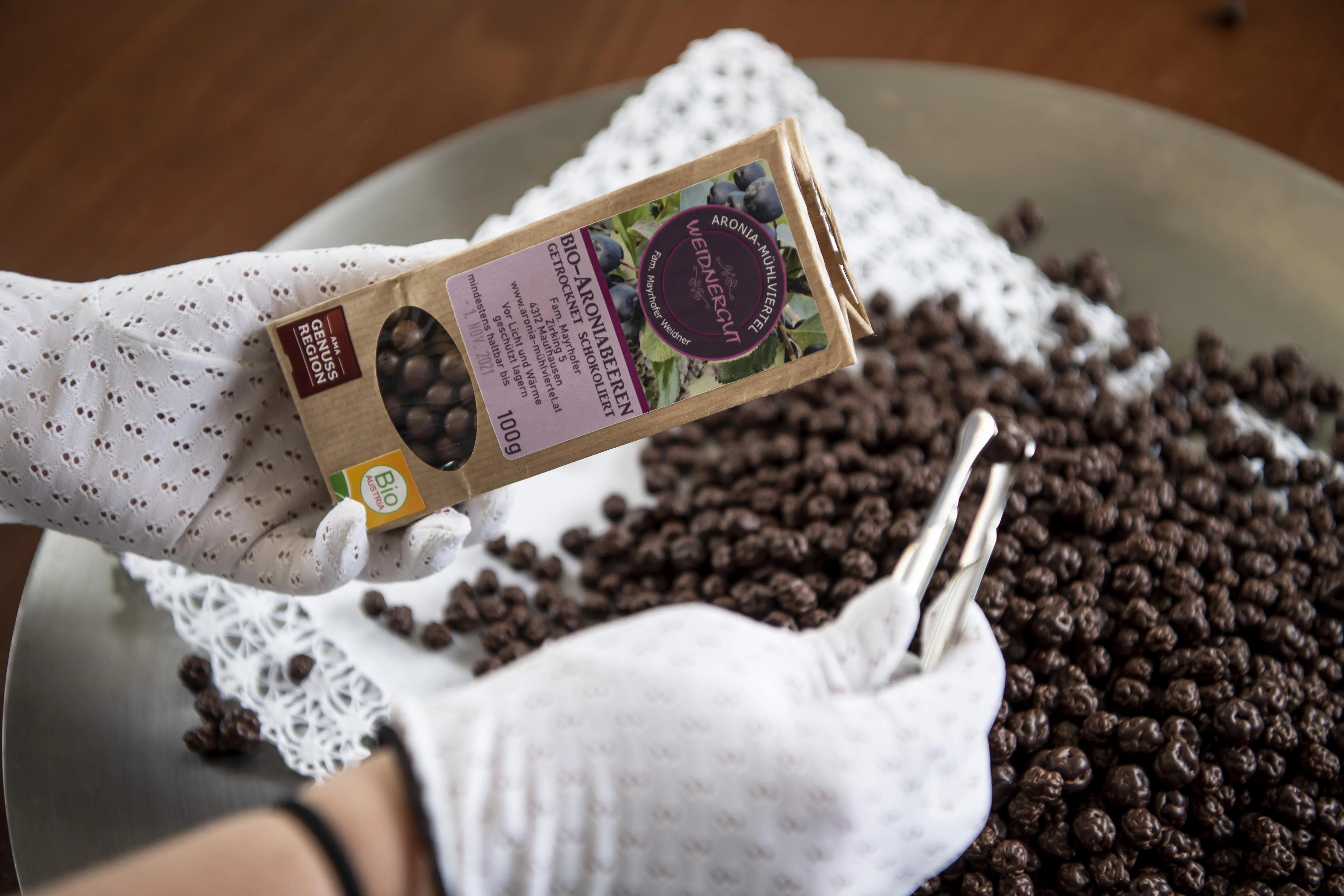 Auf dem Bild sieht man ein Teller mit weißem Deckchen, darauf schokolierte Aroniabeeren und eine Hand mit Pinzette, welche Beeren in ein Sackerl füllt.