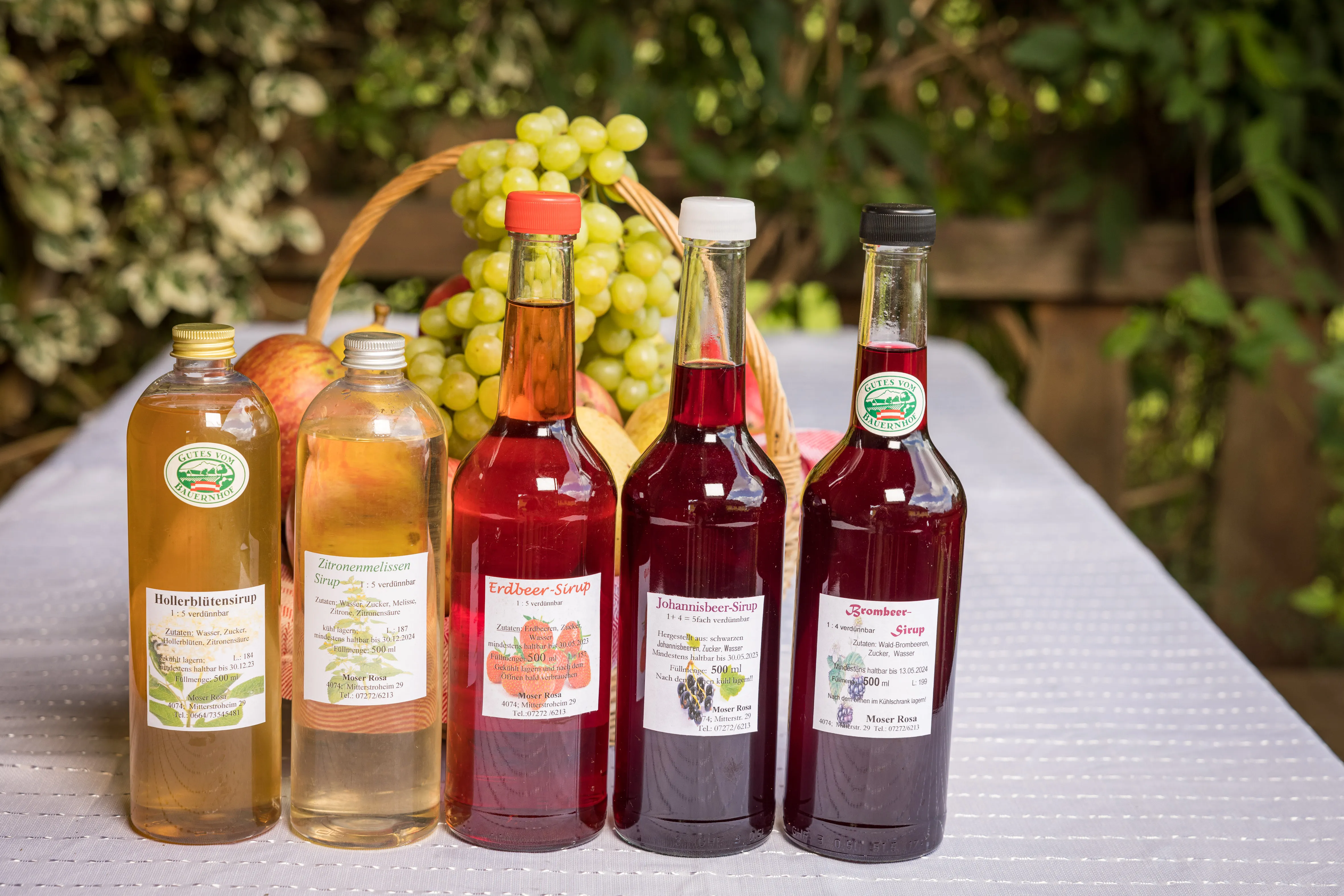 Auf einem Tisch mit weißem Tischtuch stehen fünf verschiedenfarbige Flaschen mit Sirup. Dahinter ein Korb mit Weintrauben und weiterem Obst.