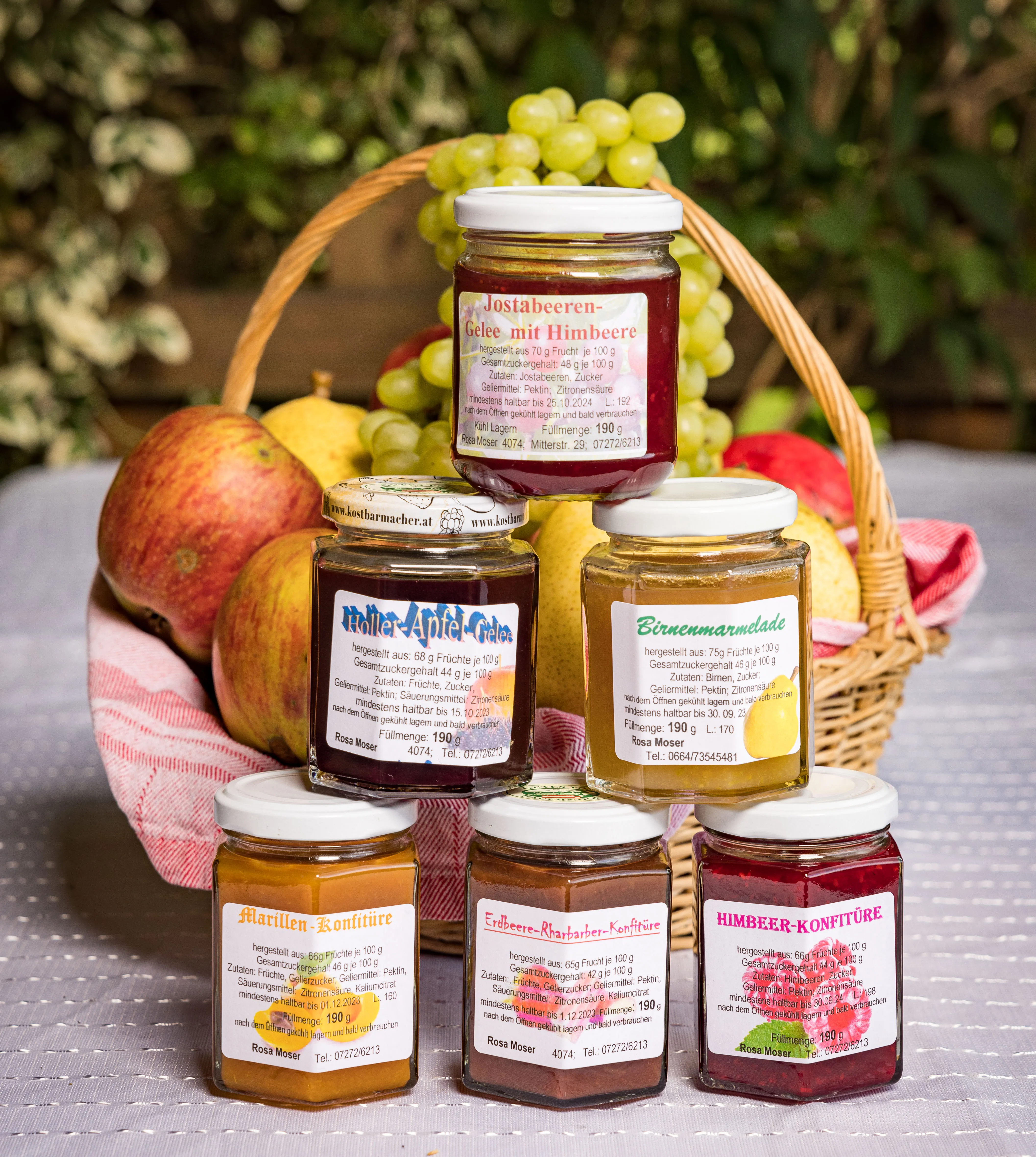 Auf einem Tisch mit weißem Tischtuch stehen verschieden farbige Gläser mit Marmelade, dahinter ein Korb mit Weintrauben und weiterem Gemüse