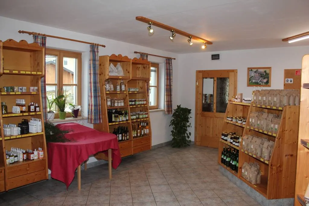 In einem weiß gestrichenen Raum stehen mehrere Holzregale an der Wand, ebenfalls ein Tisch mit rotem Tischtuch. Zwei Fenster und eine Türe sind zu sehen, bunte Vorhänge und Grünpflanzen. In den Regalen viele Produkte in Form von Flaschen, Gläsern und Fläschchen bzw. kleinen Papiersäckchen.