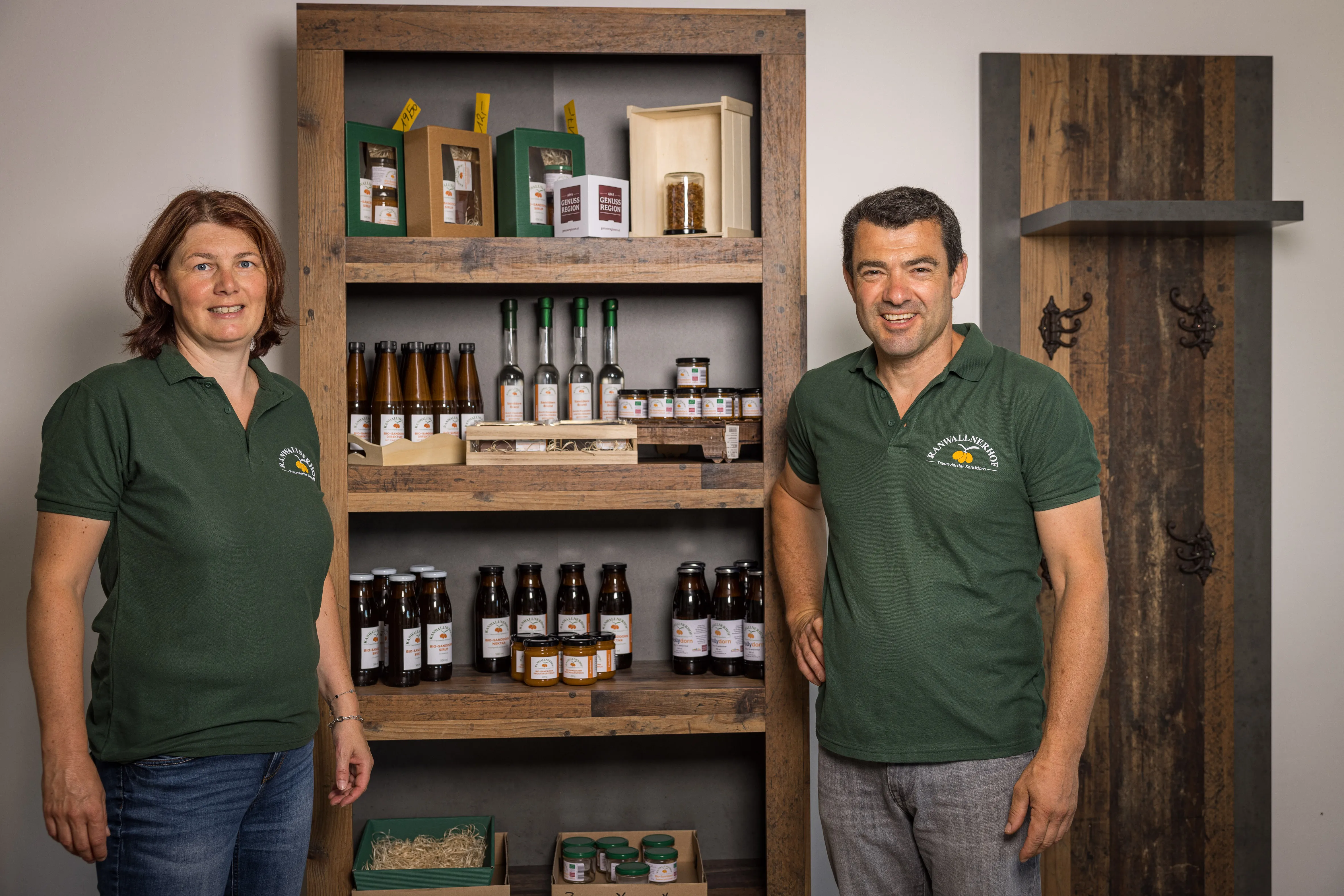 Ein Mann und eine Frau im grünen Shirt und Jeans stehen neben einem Holzregal mit verschiedenen Flaschen und Gläsern mit Sanddornprodukten