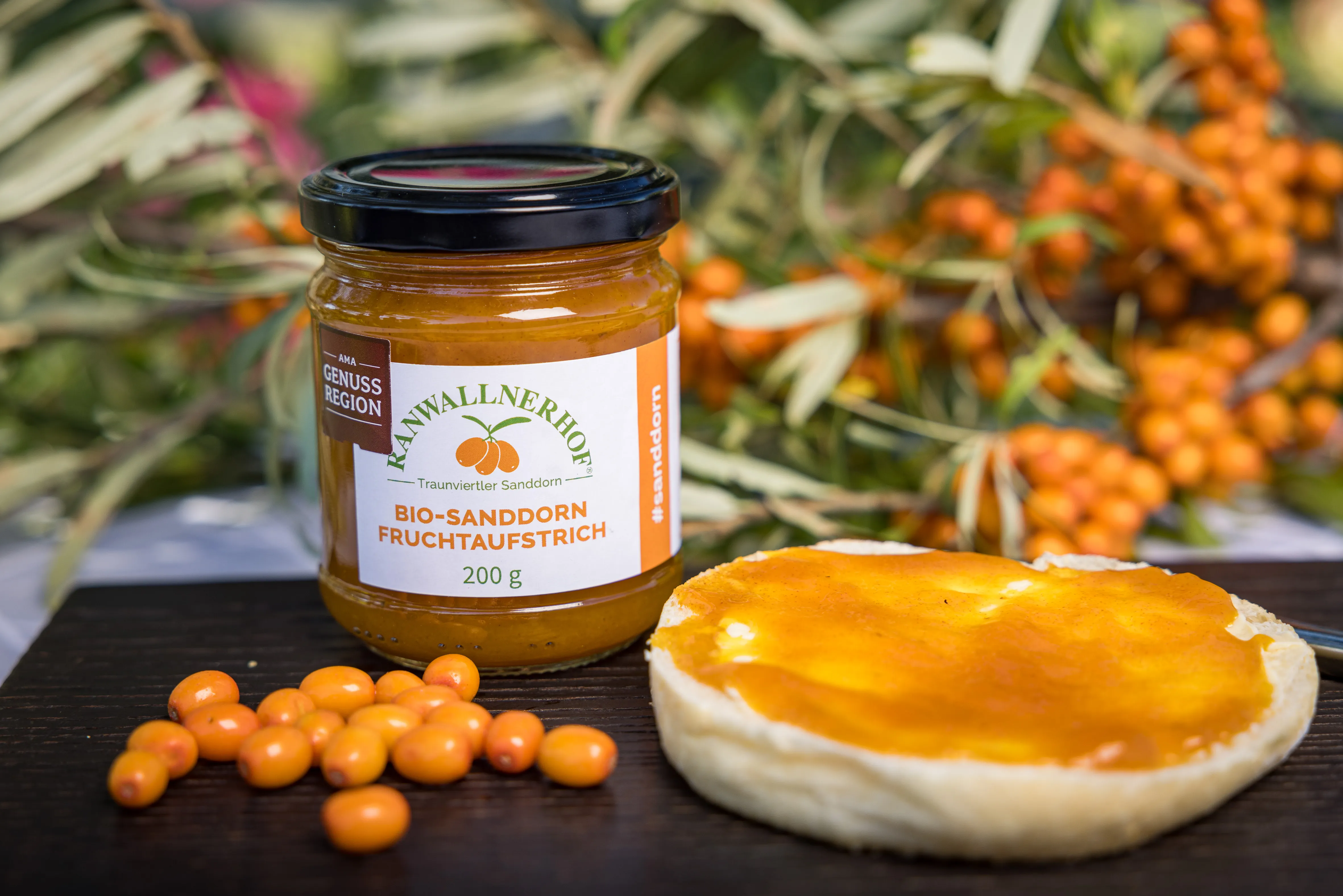 Auf einem dunklen Tisch steht ein Glas mit Sanddornaufstrich, daneben eine halbe Semmel mit Aufstrich, daneben einige orangefarbene Sanddornbeeren. Im Hintergrund Sanddornstrauch mit Früchte