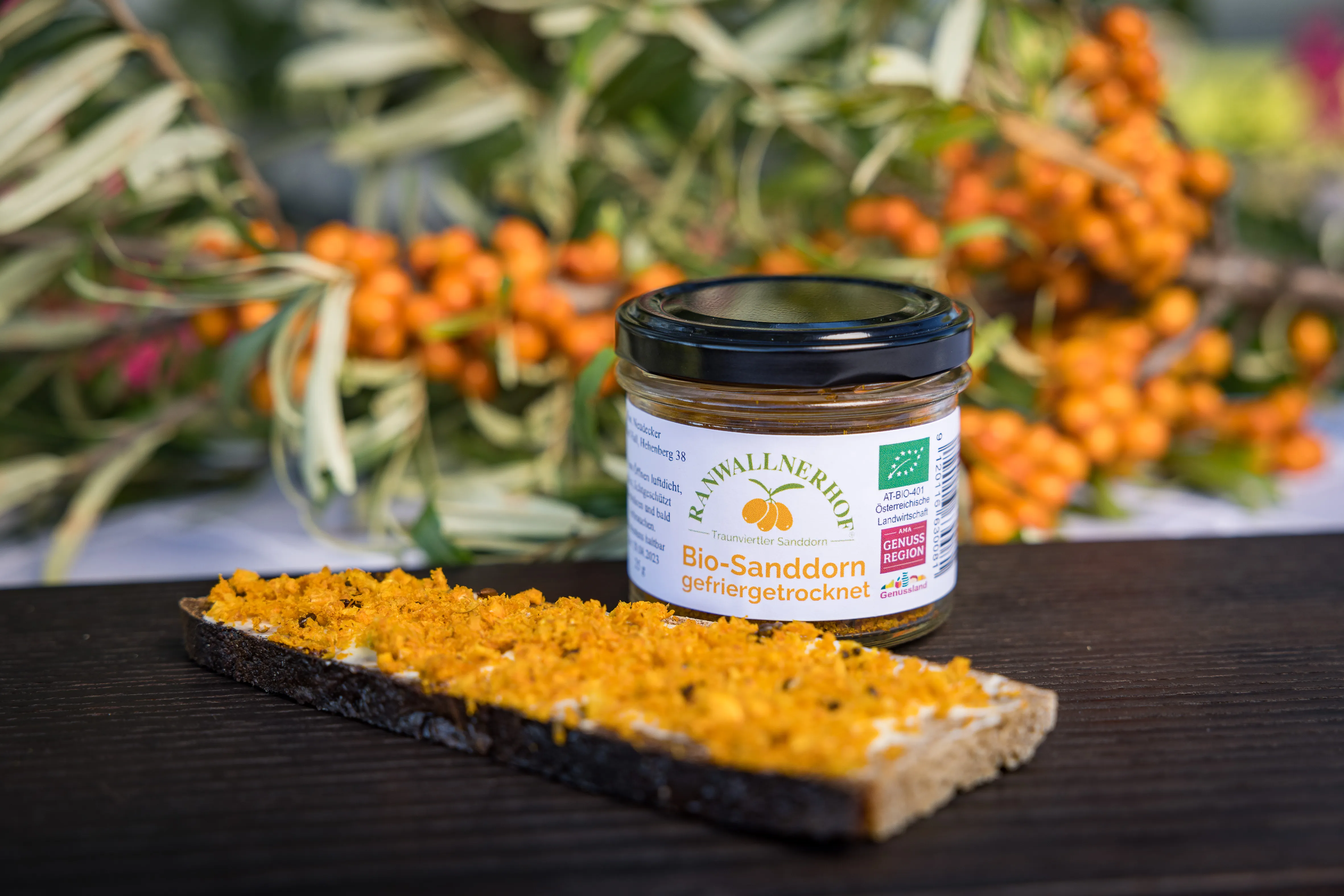 Auf einem dunklen Tisch steht ein Glas mit gefriergetrockneten Sanddornbeeren, daneben ein Butterbrot mit getrockneten Beeren. Im Hintergrund einige Sanddornäste mit Früchtenn