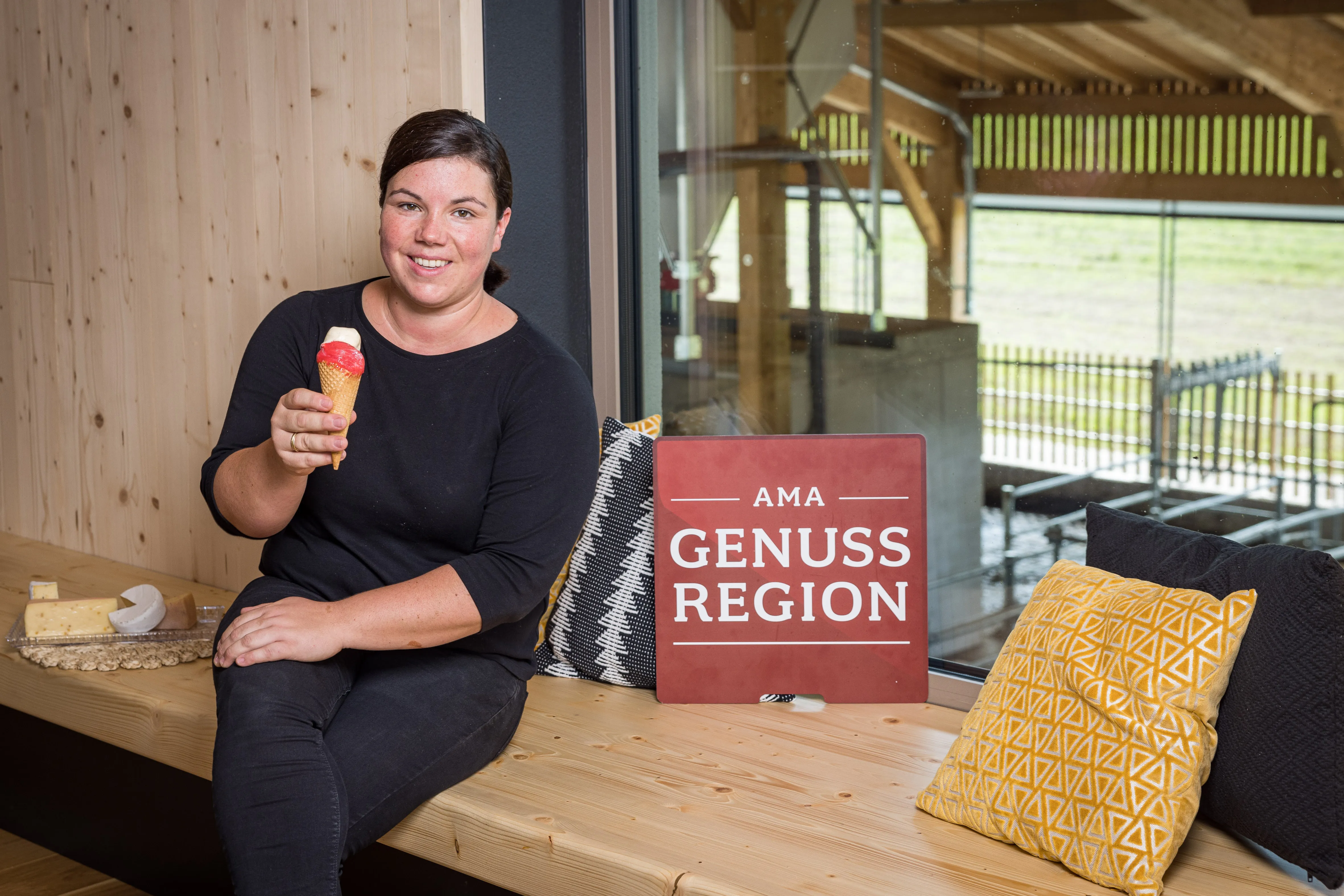 Eine dunkelhaarige Frau mit schwarzem Gewand sitzt auf einem Holztisch und hält eine Tüte mit Eis in der Hand. Daneben Ein Teller mit Käse, ein Polster, im Hintergrund der Stall