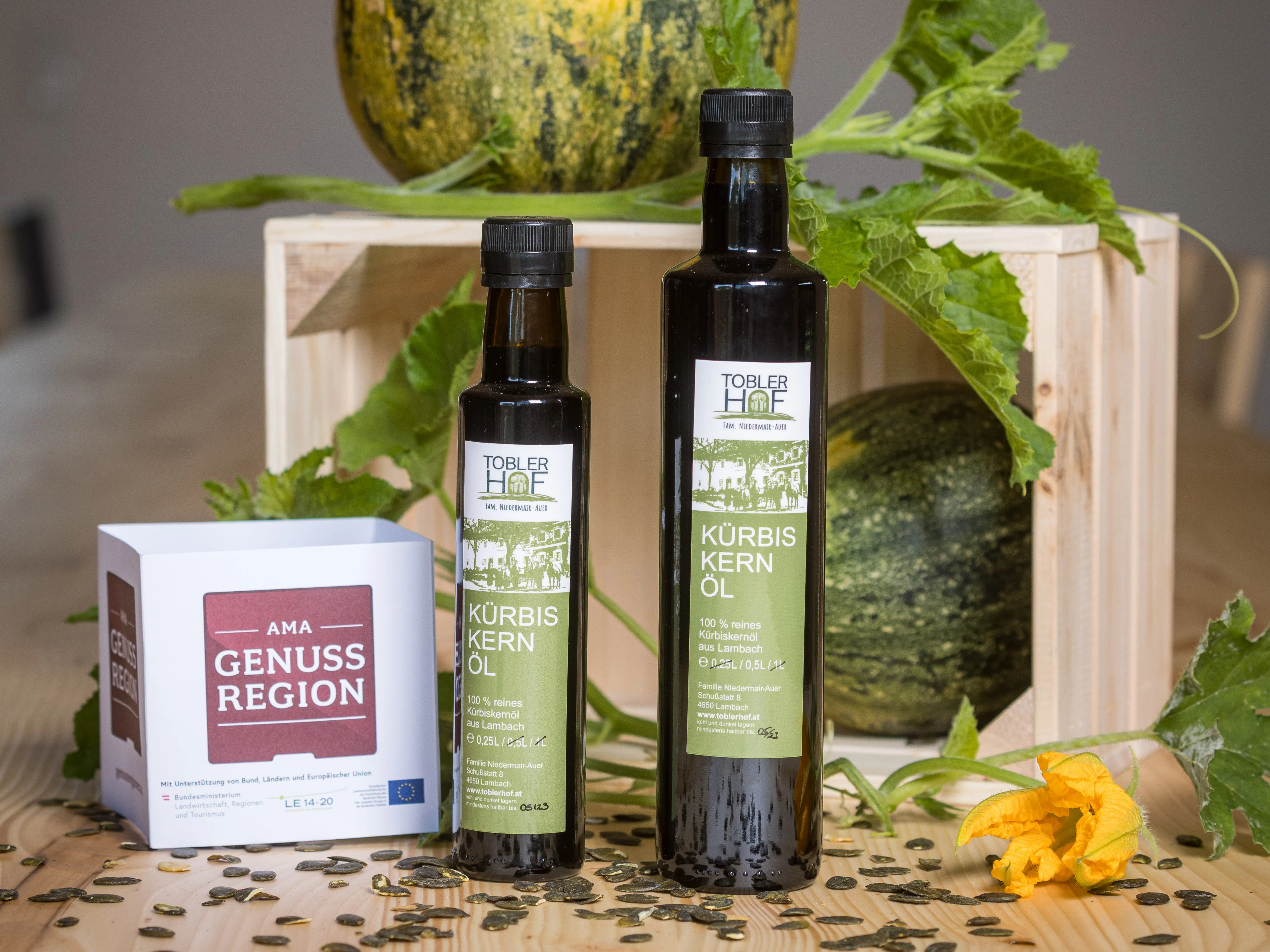 Auf einem Holztisch liegt eine Holzkiste mit einem grünen Kürbis darin und einem Kürbis darauf, davor zwei Flaschen mit Kürbiskernöl, daneben noch ein Papierwürfel mit der Aufschrift AMA Genuss Region, auf dem Tisch Kürbiskerne