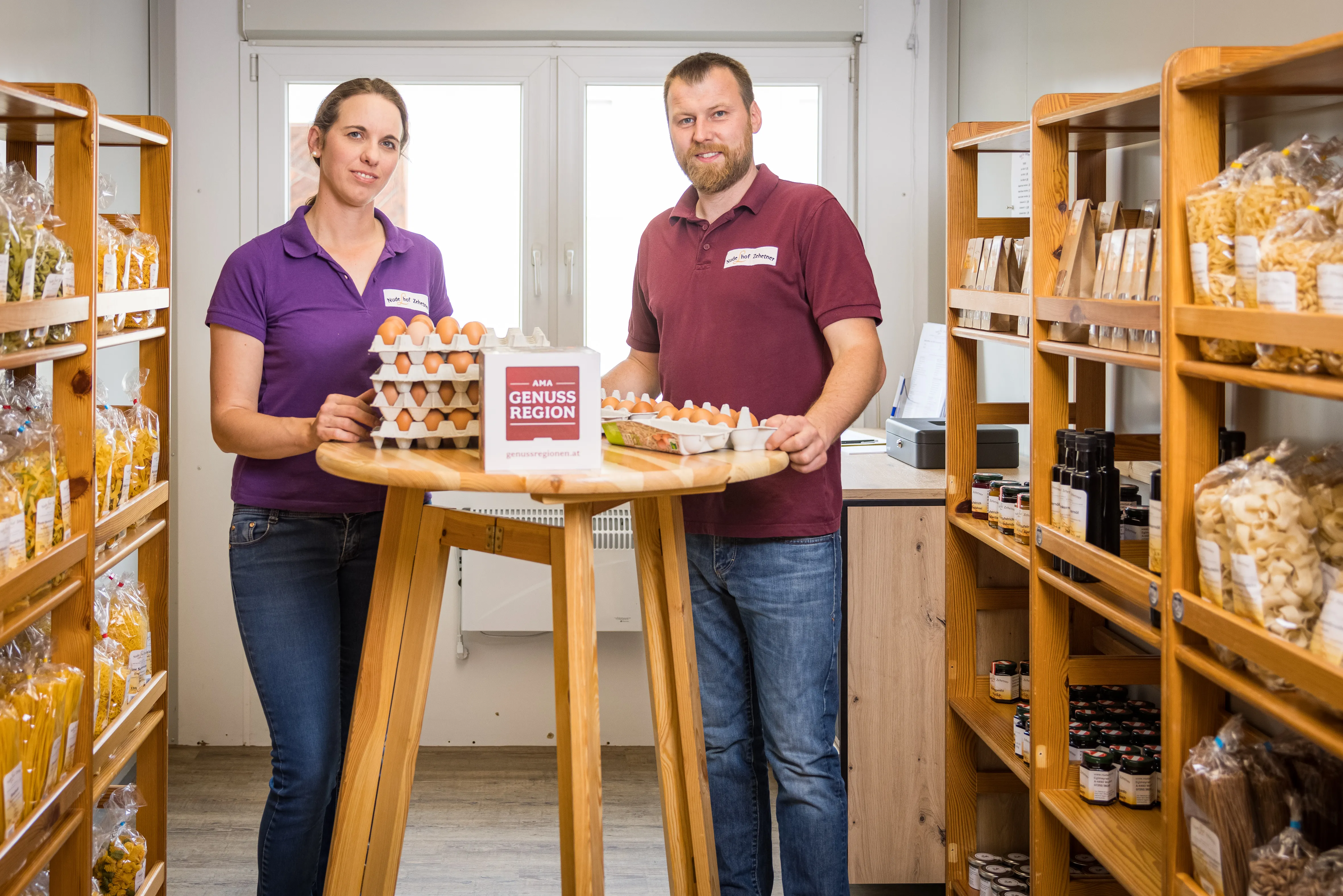 Eine Frau im lila Shirt und Jeans und ein Mann im roten Shirt und Jeans stehen an einem Stehtisch in einem Verkaufsladen. Auf dem Stehtisch stehen mehrere Eierhöcker mit braunen Eiern und eine Tafel mit AMA GENUSS REGION. Daneben stehen Regale an der Wand mit verschiedenen Nudelpackungen, Flaschen und anderen Verpackungen.
