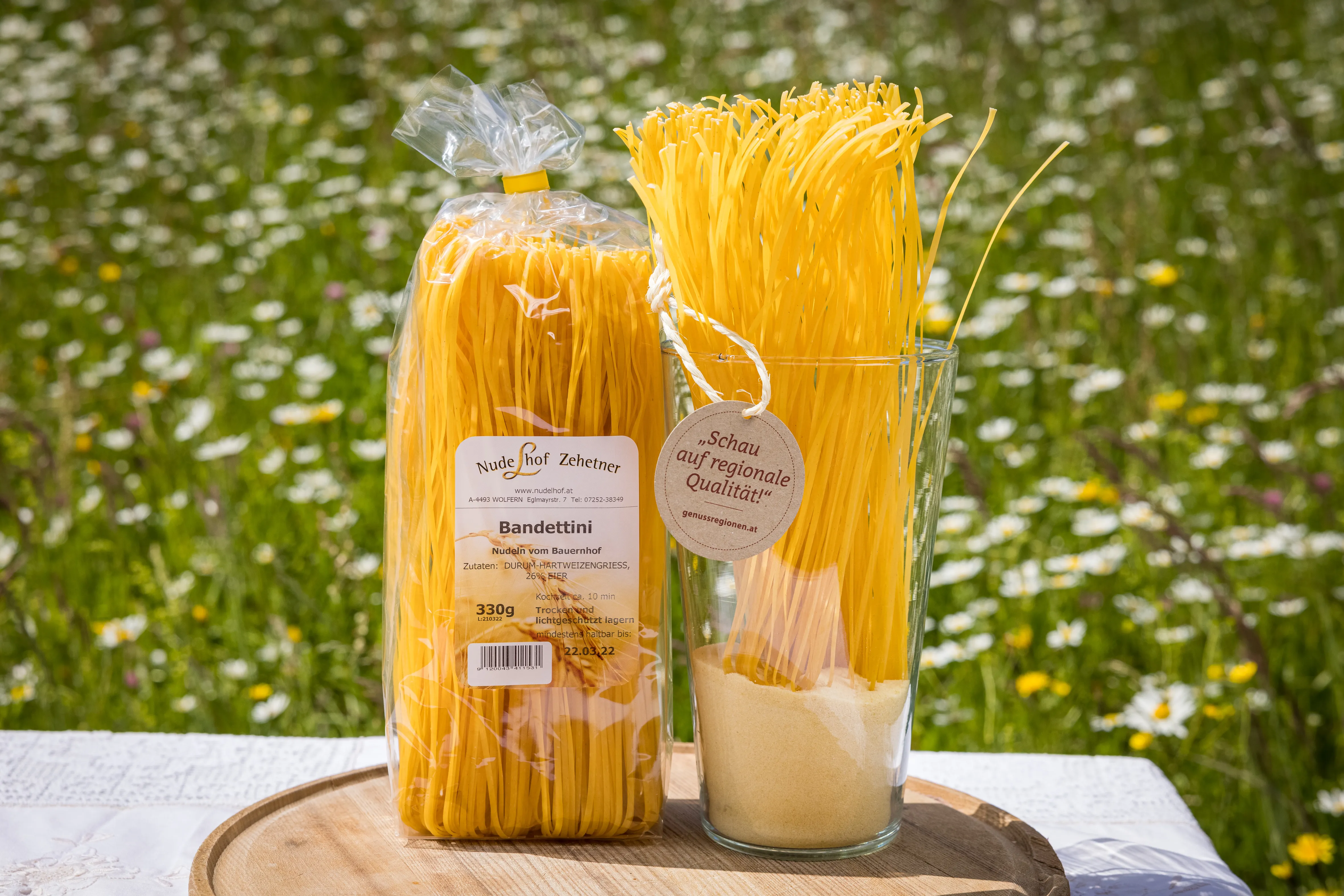 Auf einem hellen Tisch liegt ein Holzbrett mit zwei Nudelpackungen mit gelben Spaghetti. Dahinter eine grüne Wiese mit vielen Margeriten und Löwenzahn.