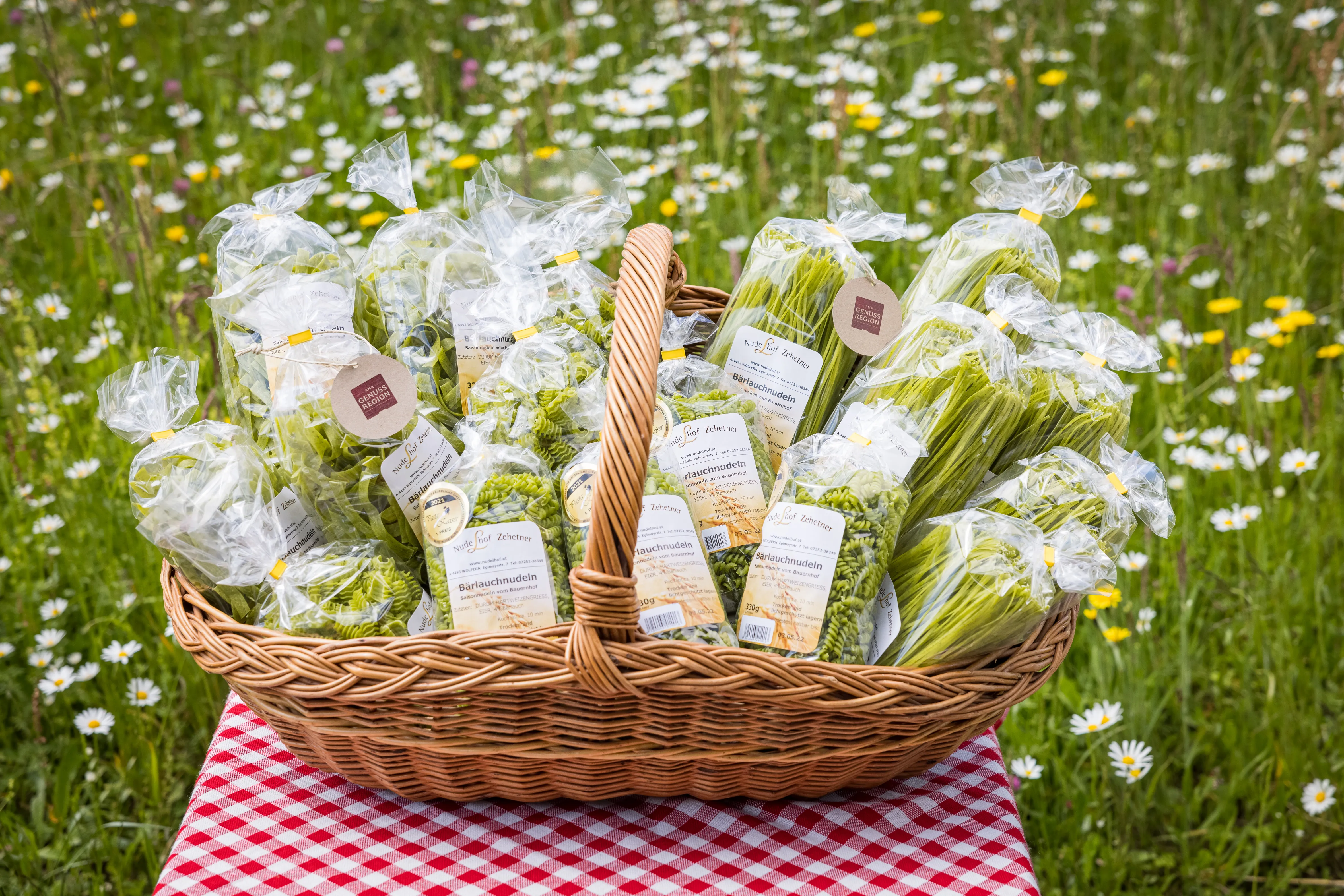 Ein brauner Korb mit vielen verschiedenen Nudelpackungen mit grünen Nudeln steht auf einem Tisch mit einem weiß/rot karierten Tischtuch. Dahinter grüne Wiese mit weißen Margeriten und Löwenzahn.