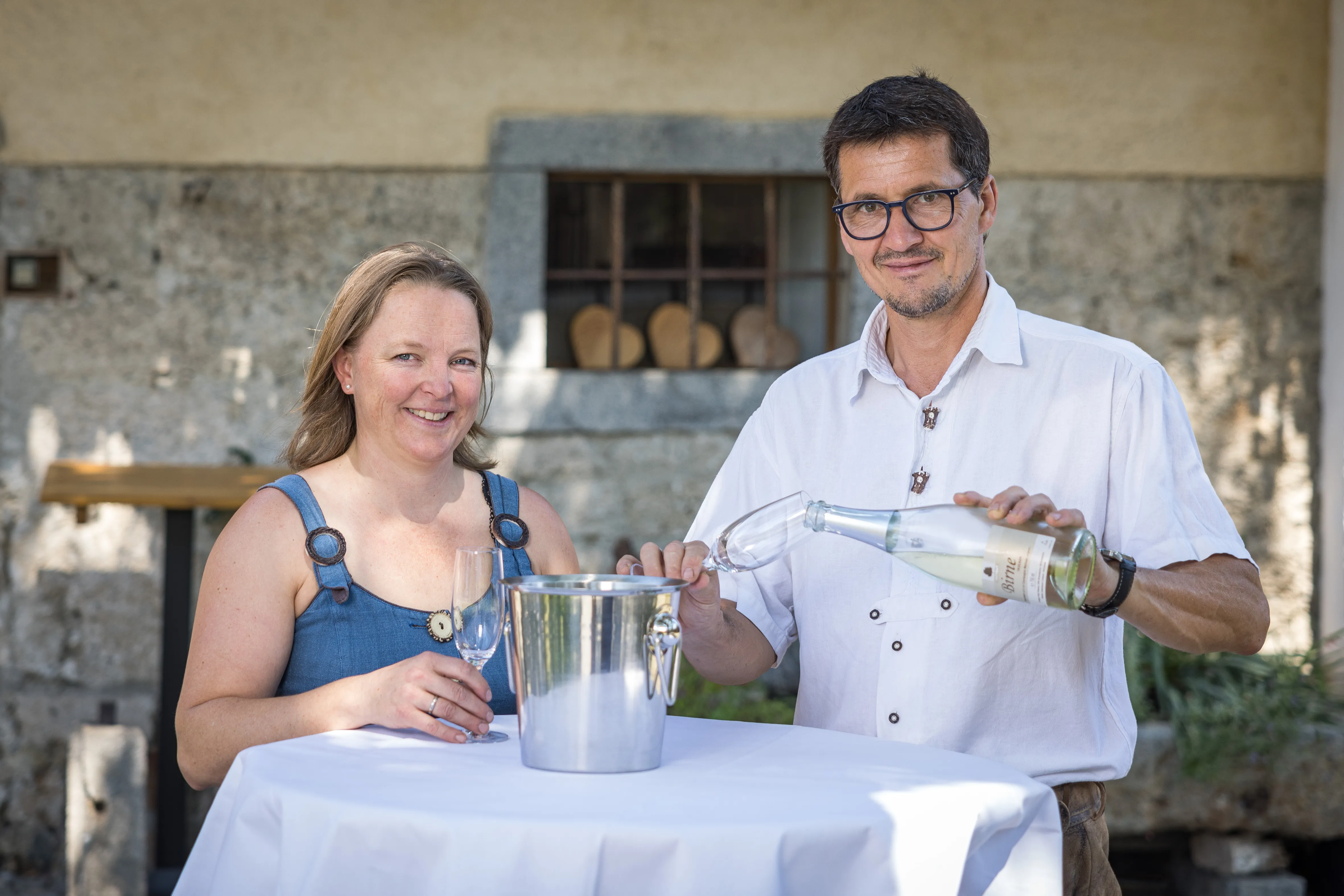 Herr und Frau Ober stehen an eine Stehtisch mit weißen Tischtuch, darauf steht ein Sektkühler. Frau Ober hält ein Glas und Herr Ober schenk aus einer Flasche in ein zweites Glas ein. 