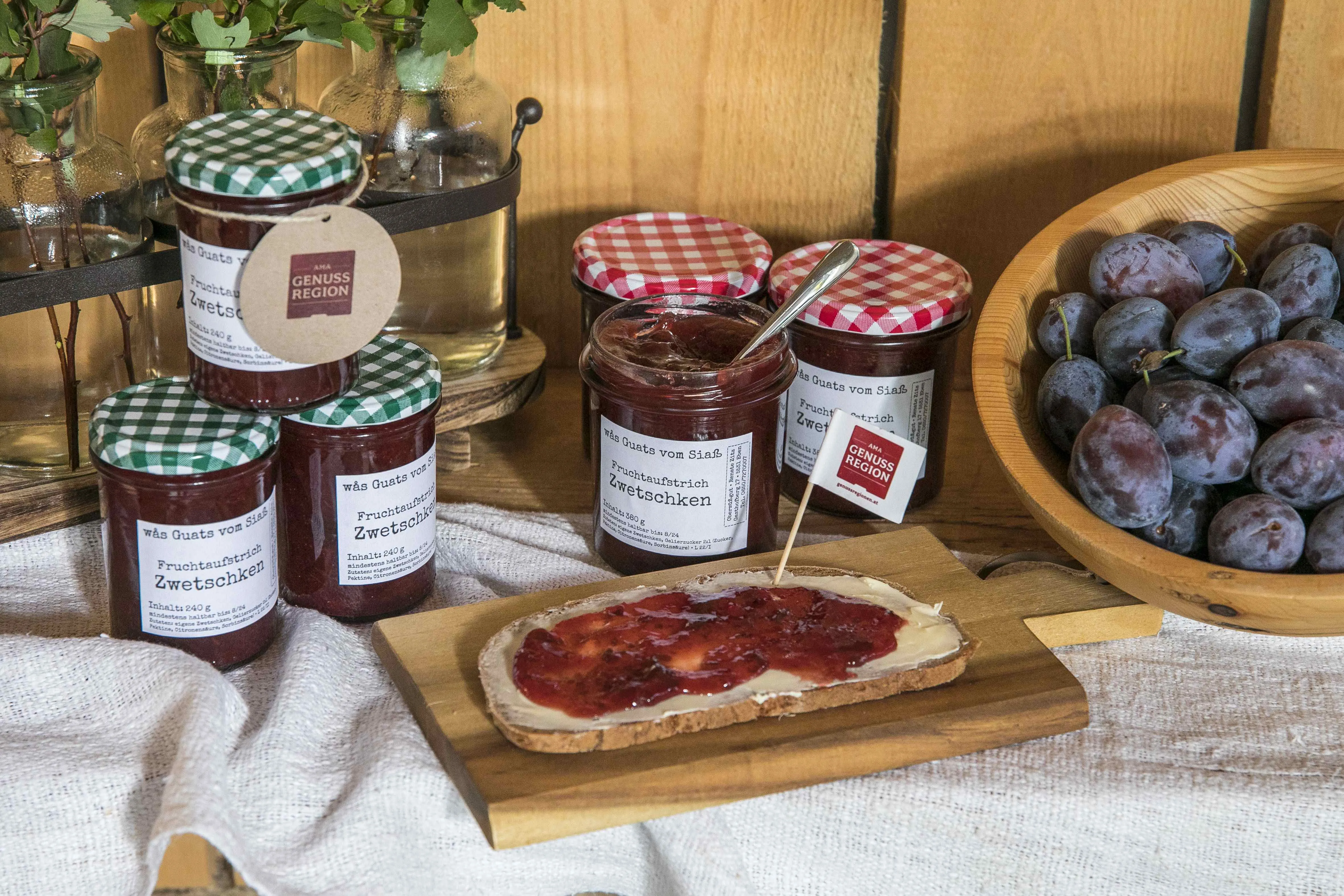 Fruchtaufstriche der Fam. Zitz links im Bild 3 Stk mit grün kariertem Deckel gestapelt, mittig ein aufgestrichenes Brot und dahinter drei Gläser mit rot kartierten Deckeln, daneben Zwetschgen in einer Holzschale