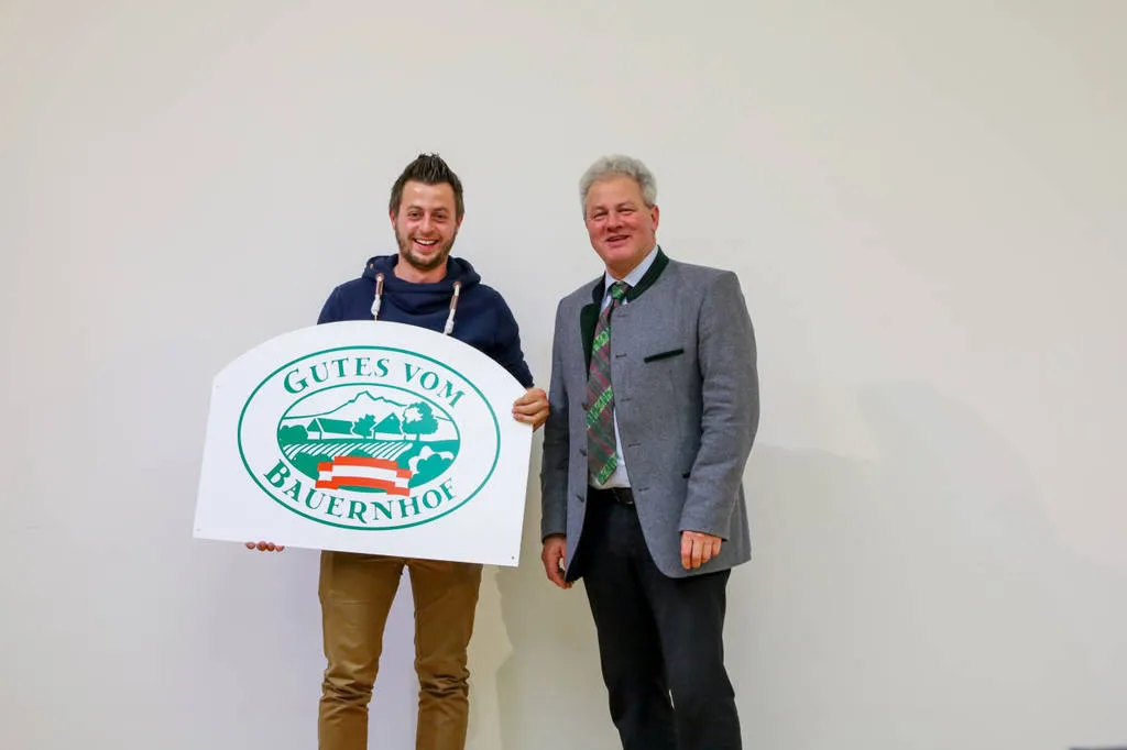 Herr Ortner und ein weiterer Mann stehen vor einem weißen Hintergrund und halten eine Hoftafel in den Händen