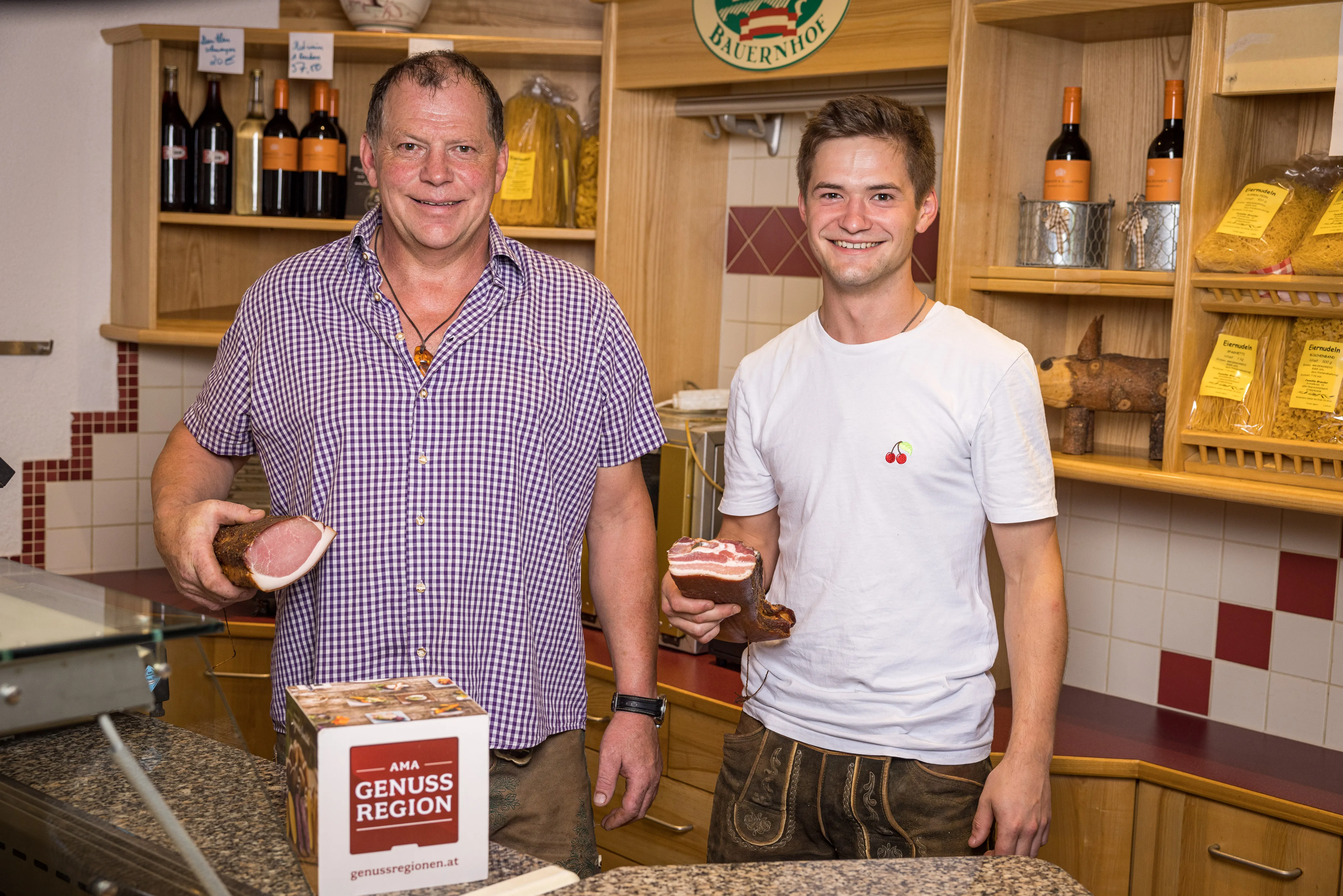 Herr Peneder sen. und jun. stehen in einem Hofladen und halten jeweils ein Stück Speck in der Hand. vor Ihnen eine Theke mit Waage, dahinter Holzregale mit verschiedenen Produkten