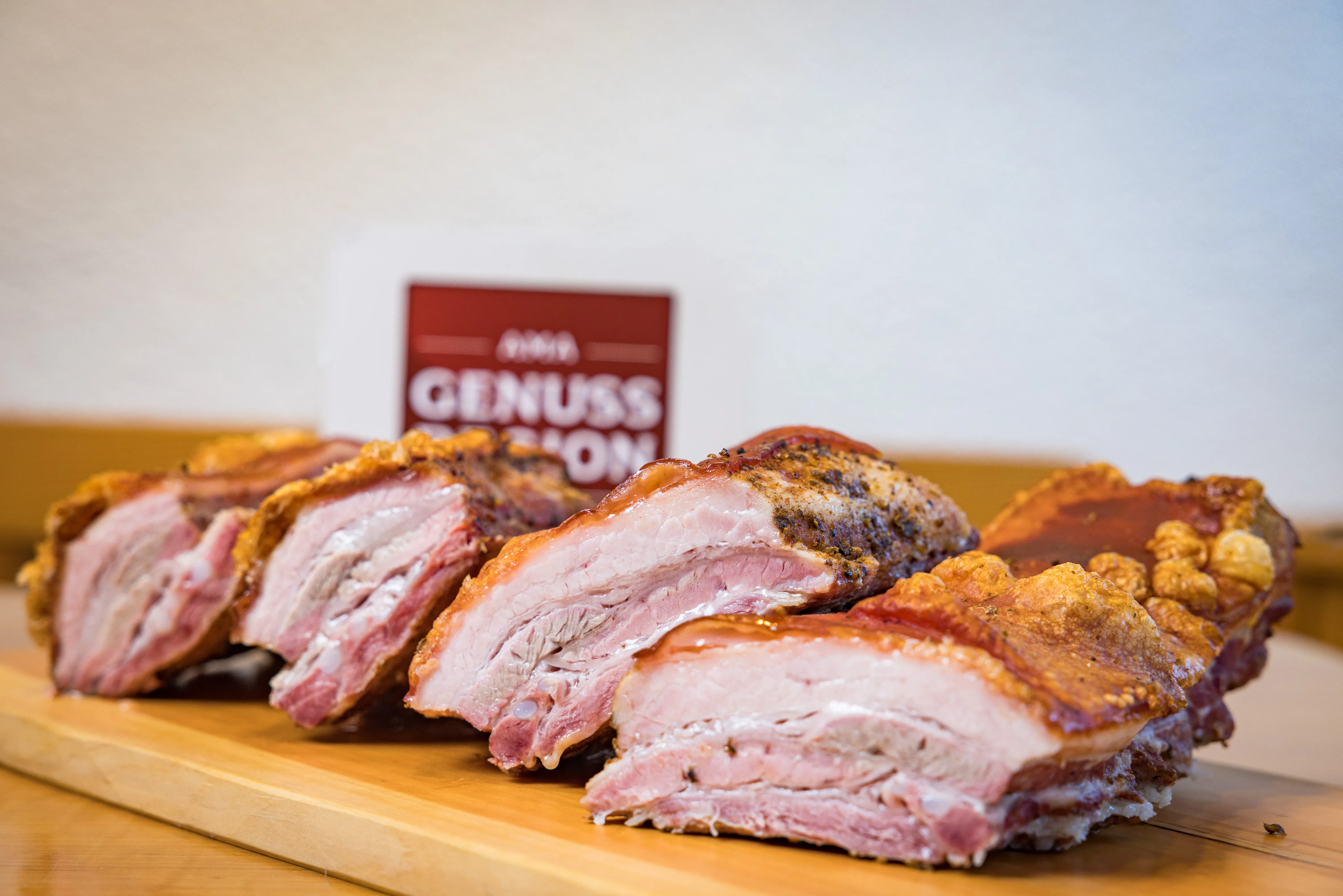 Auf einem Holztisch liegen aufgeschnittene Bratenstücke mit Kruste, dahinter weiße Wand mit Ama Genuss Tafel