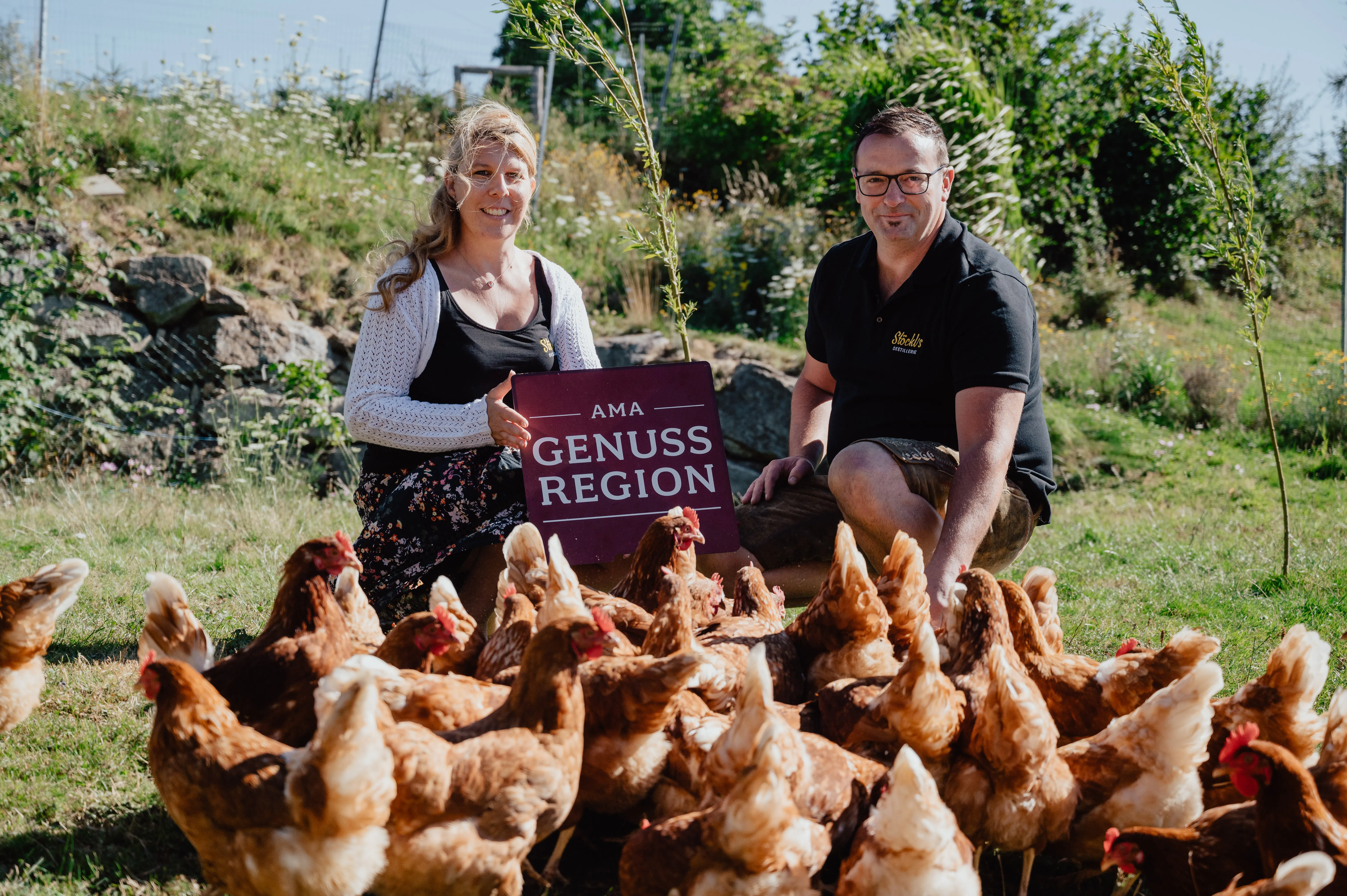 Ein Mann und eine Frau knien auf einer grünen Wiese, ein Schild mit der Aufschrift AMA GENUSS REGION in den Händen; davor viele braune Hühner. Im Hintergrund Büsche und Himmel