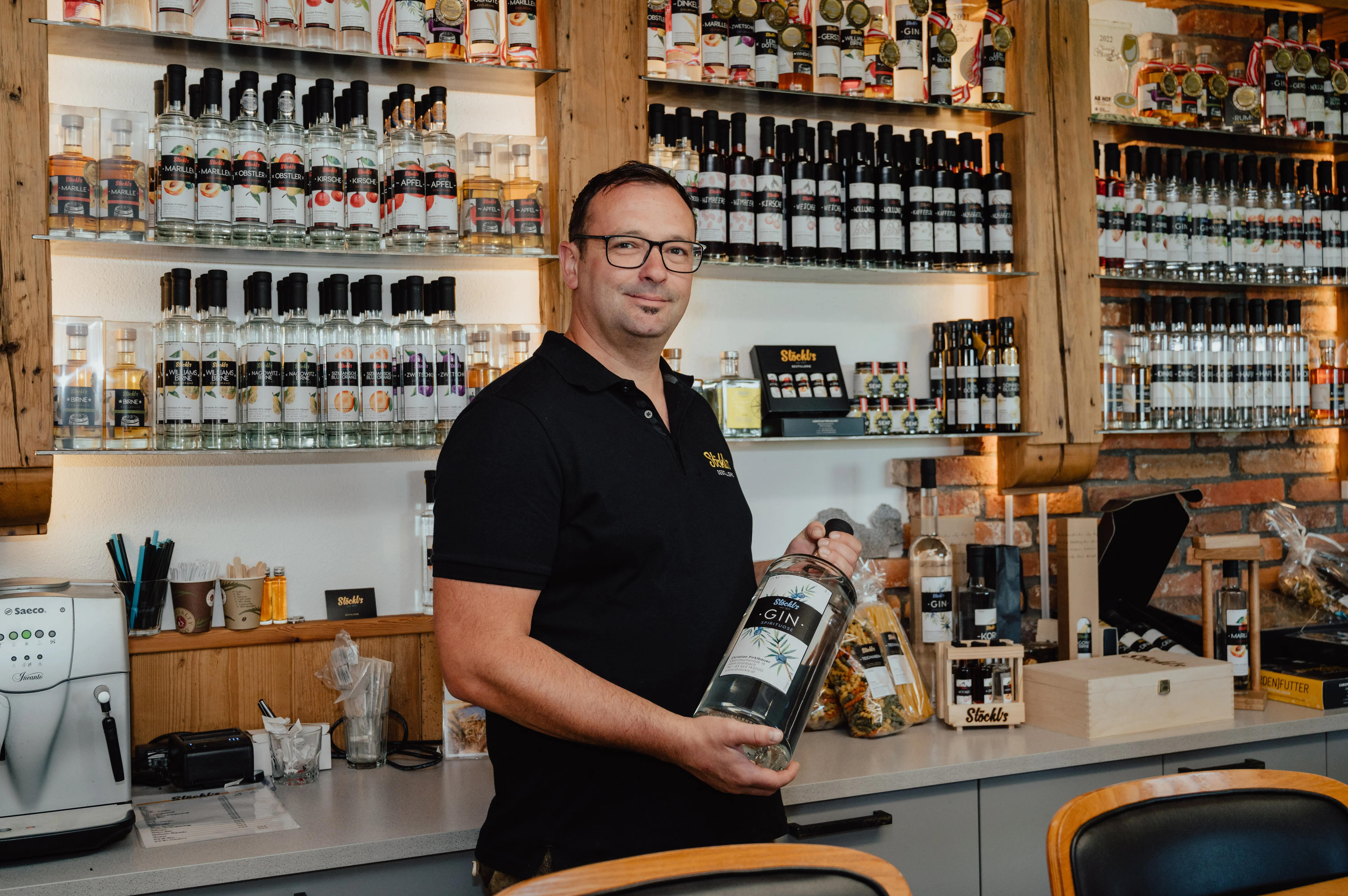 Herr Pirklbauer steht vor einem großen Regal mit verschiedenen Flaschen und Produkten. Er hält eine Flasche in den Händen. Dahinter eine Anrichte mit Schubladen.