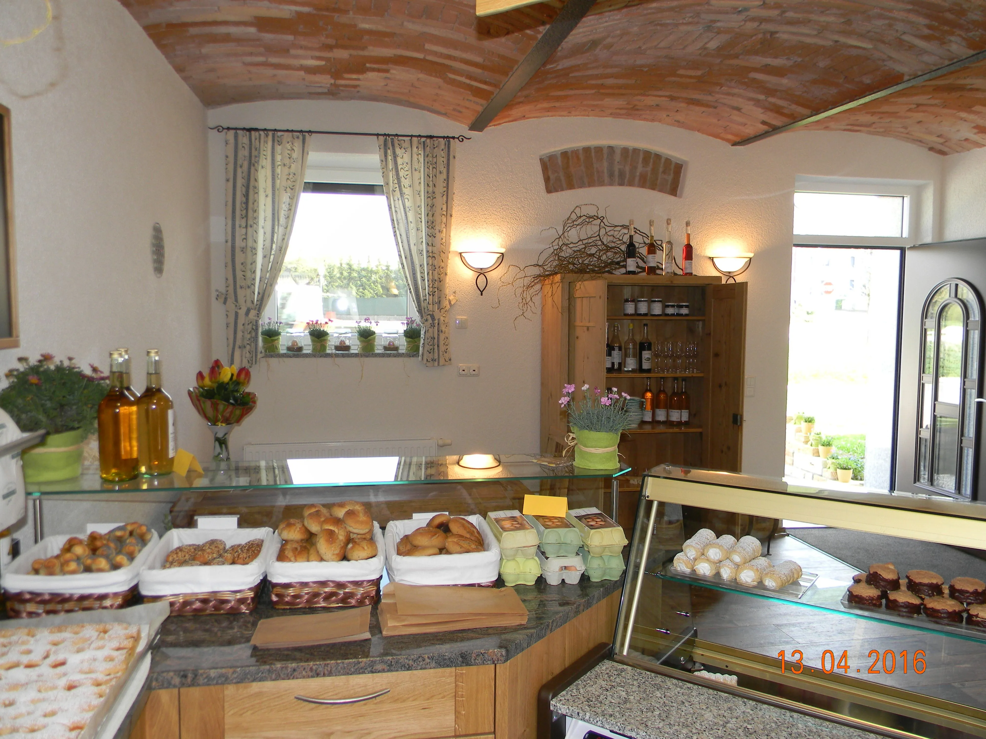 In einem weiß gestrichenen Raum befinden sich eine Theke mit verschiedenen Produkten, wie Eier, Brot und Gebäck. Man sieht ein Fenster und eine Tür, an der Wand ein weiterer Schrank mit Produkten.