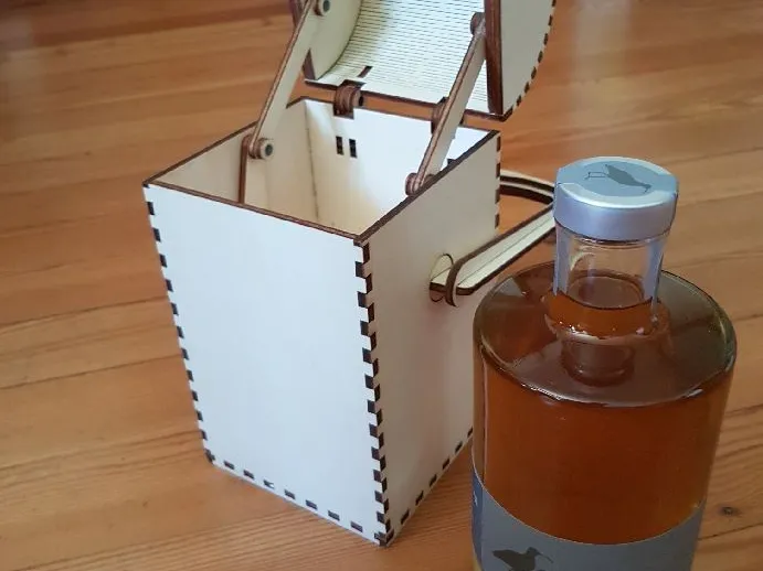 Auf einem Tisch steht eine Flasche mit dunkelgelbem Inhalt, daneben eine weiße Verpackung in Form einer Schatzkiste