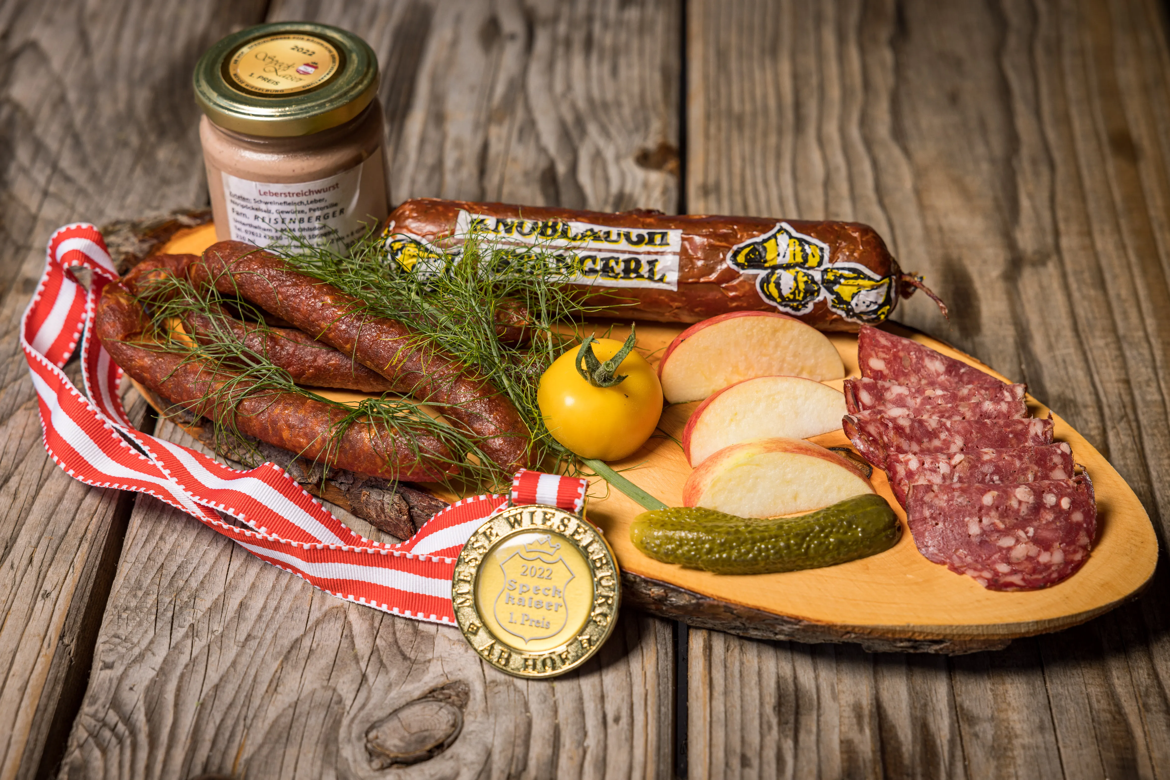 Auf einem Holztisch liegt ein großes ovale Jausenbrett mit ganzer und aufgeschnittener Wurst. garniert mit Äpfel, Gurkerl und gelber Tomate. Daneben noch ein Glas mit Senf.